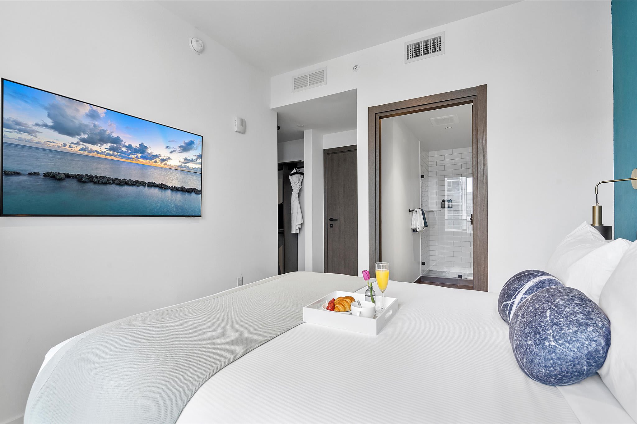A modern bedroom featuring a queen-size bed with a light upholstered headboard, a wall-mounted flat-screen TV, and an open doorway leading to an ensuite bathroom. A breakfast tray rests on the bed.