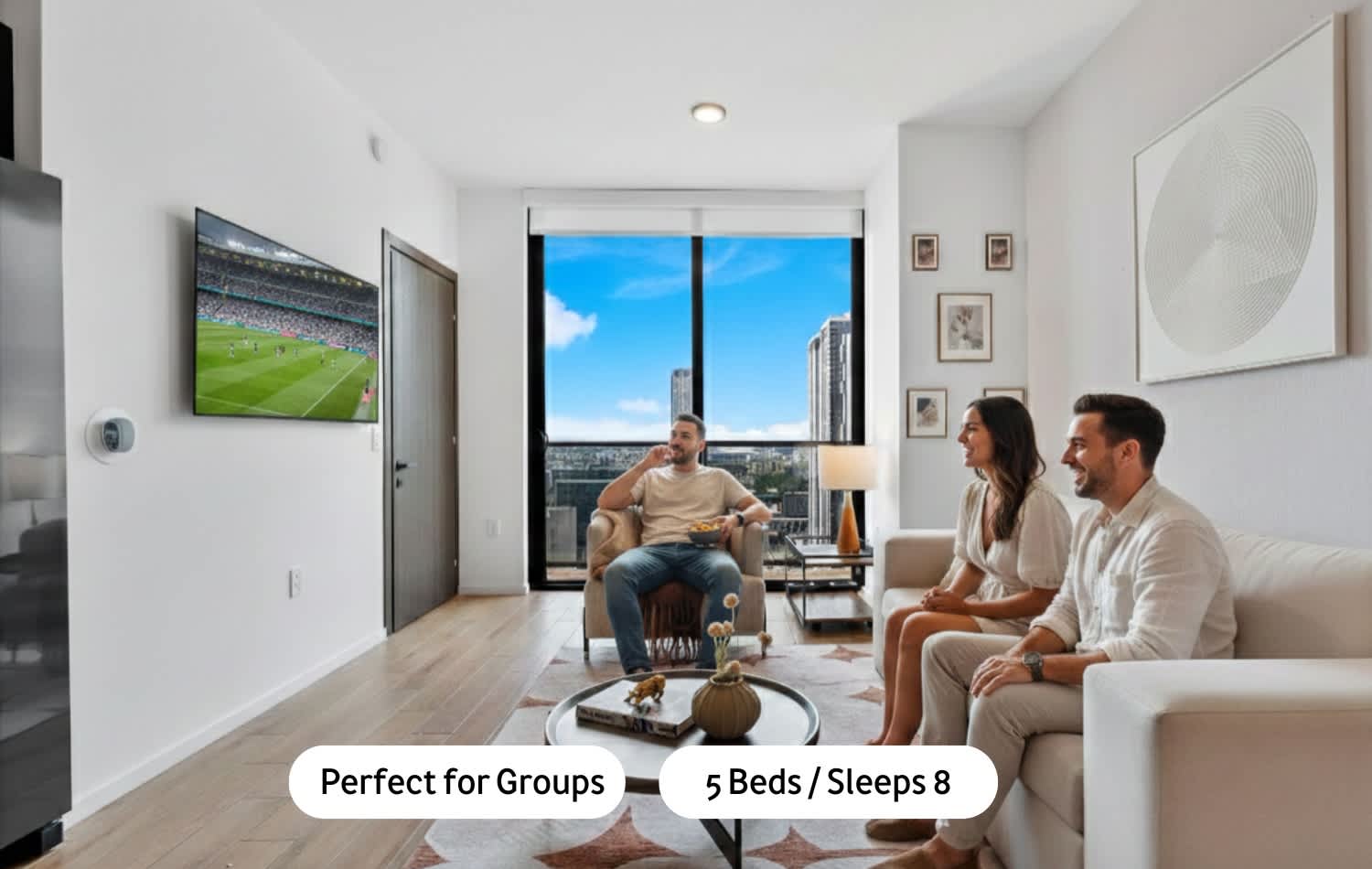 Modern living room with light-colored walls and wood flooring, featuring a large wall-mounted TV, a comfortable sofa, and an armchair, all facing a large window with a city skyline view. Three people are relaxing in the space.