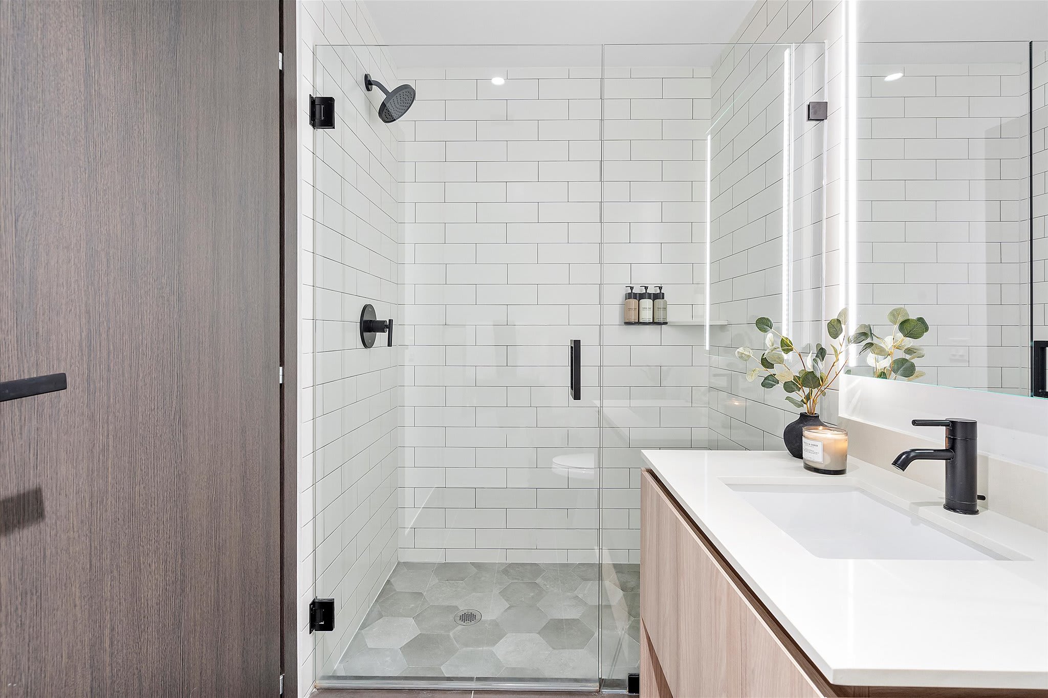 A modern bathroom with a walk-in shower featuring white subway tiles and a hexagonal gray floor, alongside a light wood vanity with a white countertop and a large illuminated mirror. Black fixtures add a contemporary touch.