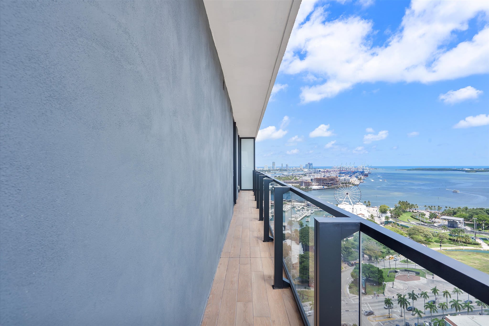Long, narrow balcony with light wood-look tile flooring and a glass railing, offering a panoramic view of a bay with boats, a Ferris wheel, and distant cityscapes under a blue sky. A gray textured wall runs along one side.