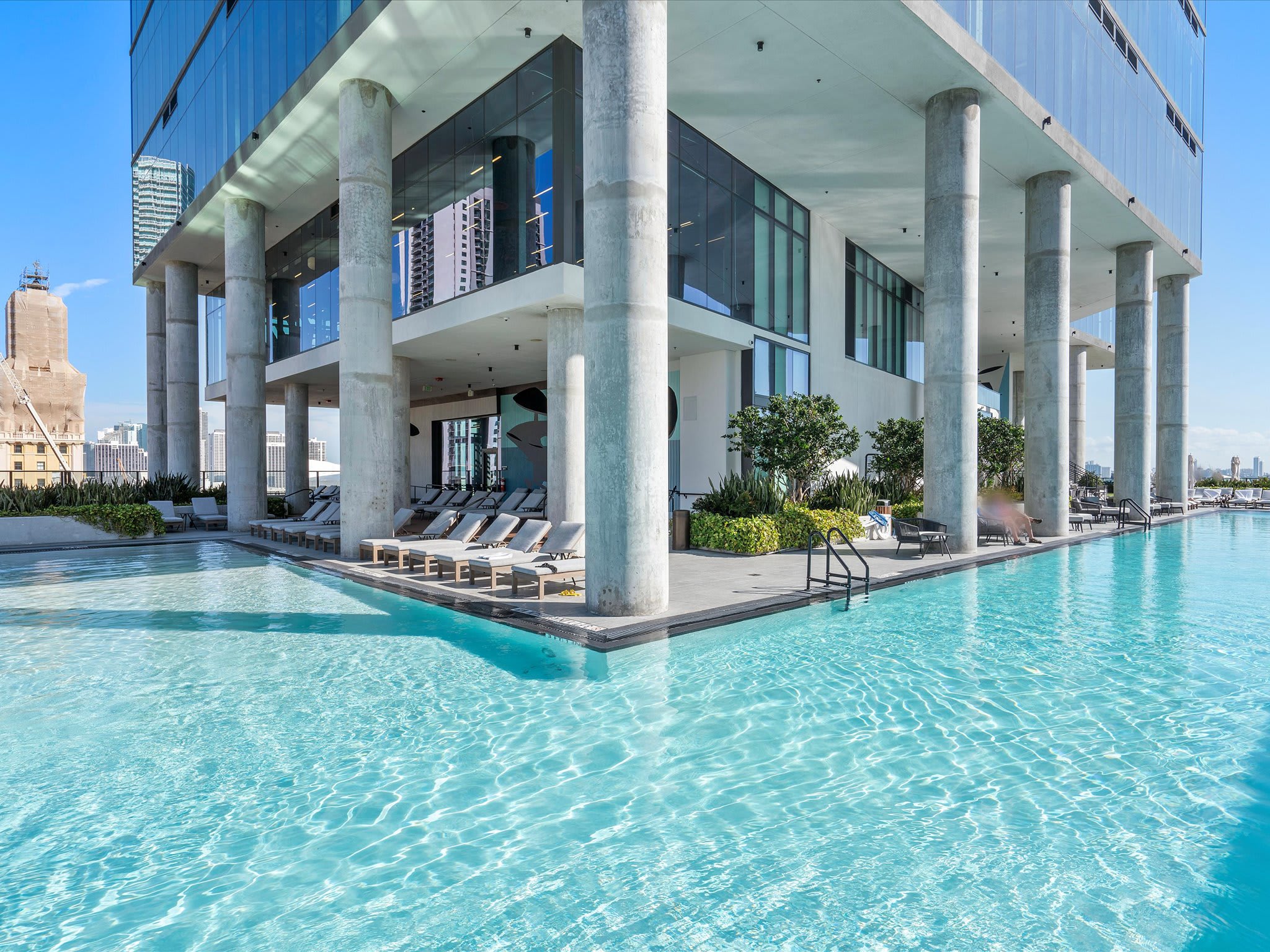 Large, sparkling blue swimming pool surrounded by lounge chairs and a modern building with large glass windows and concrete pillars. The pool area offers a luxurious communal space for relaxation.