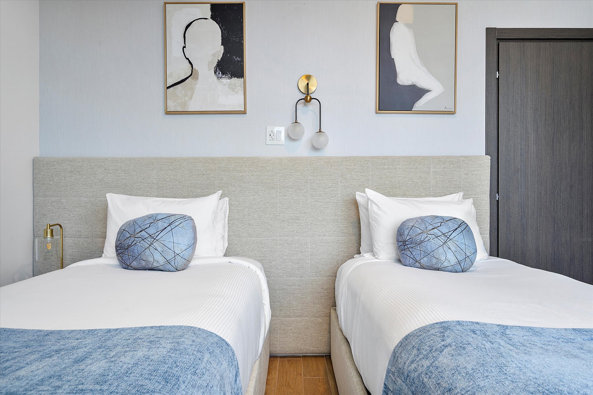 A close-up view of two twin beds with light upholstered headboards, crisp white linens, and decorative blue pillows. Two abstract artworks hang above the beds, complemented by modern wall sconces.