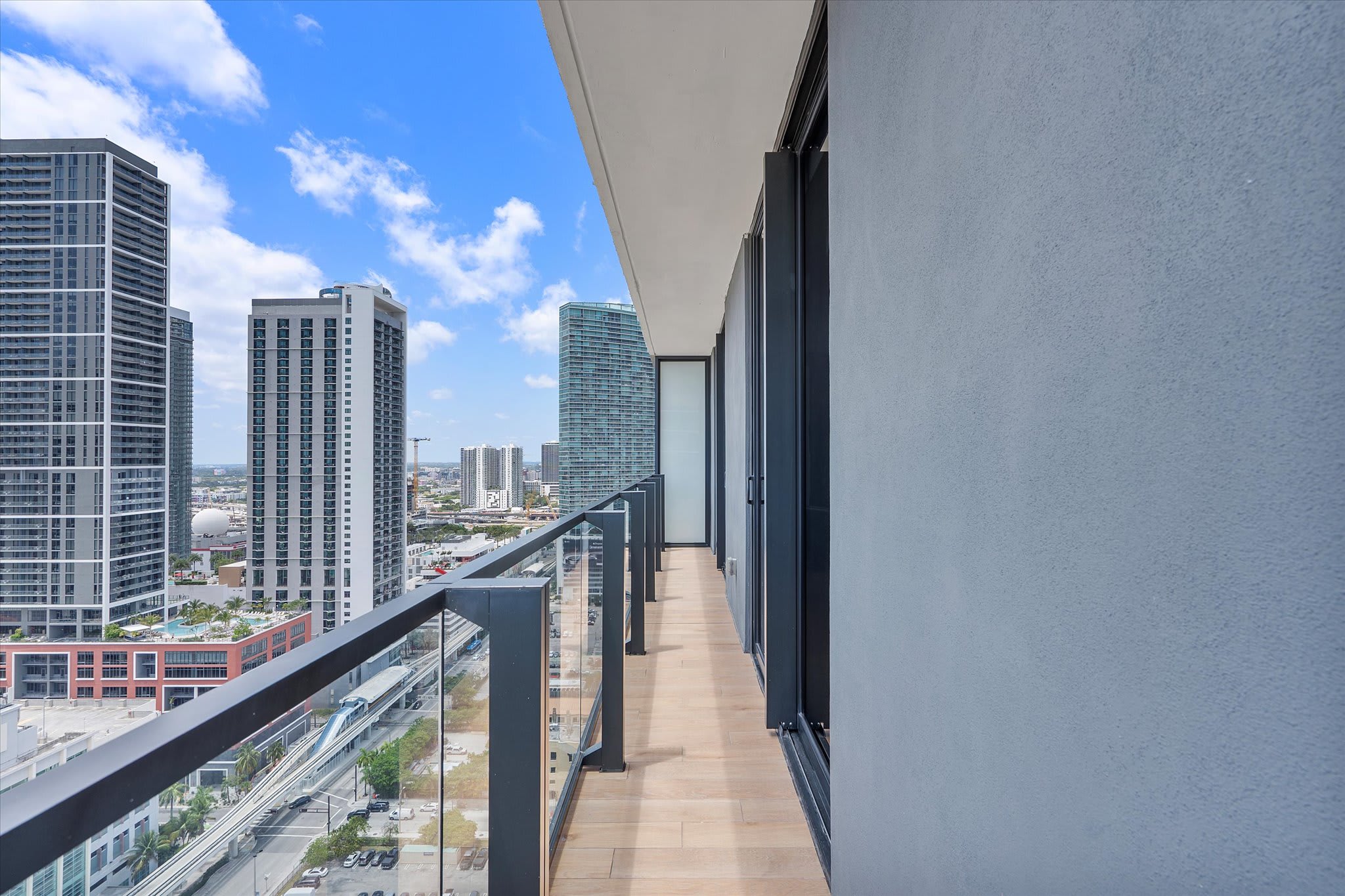 Expansive balcony with light wood-look tile flooring and a glass railing, providing a sweeping view of modern high-rise buildings and a city train line. The sky is bright blue with scattered clouds.