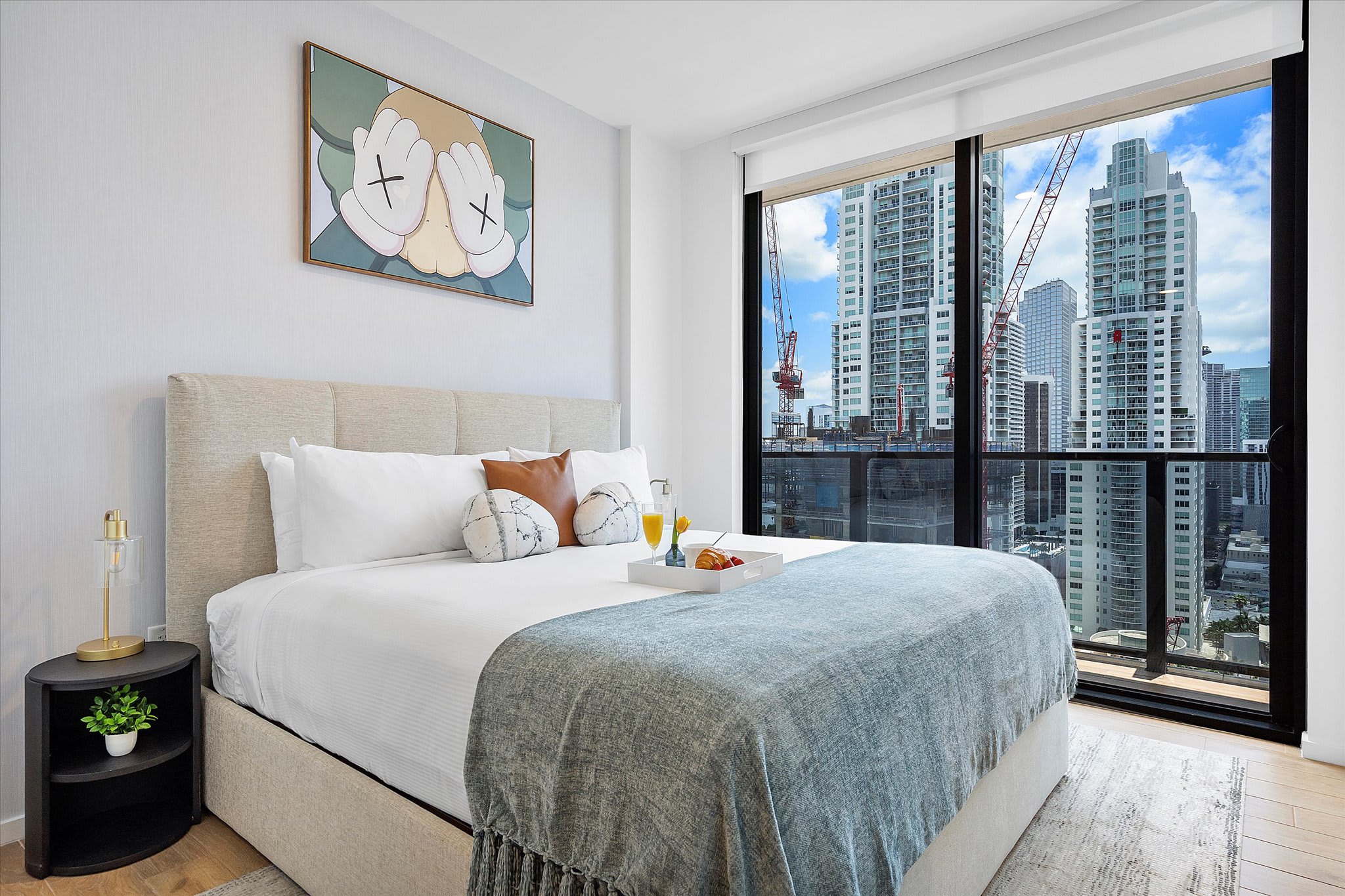 A spacious bedroom with a light upholstered king-size bed, a breakfast tray with food, and floor-to-ceiling windows offering panoramic city views with construction cranes. A large KAWS-style artwork hangs above the bed.