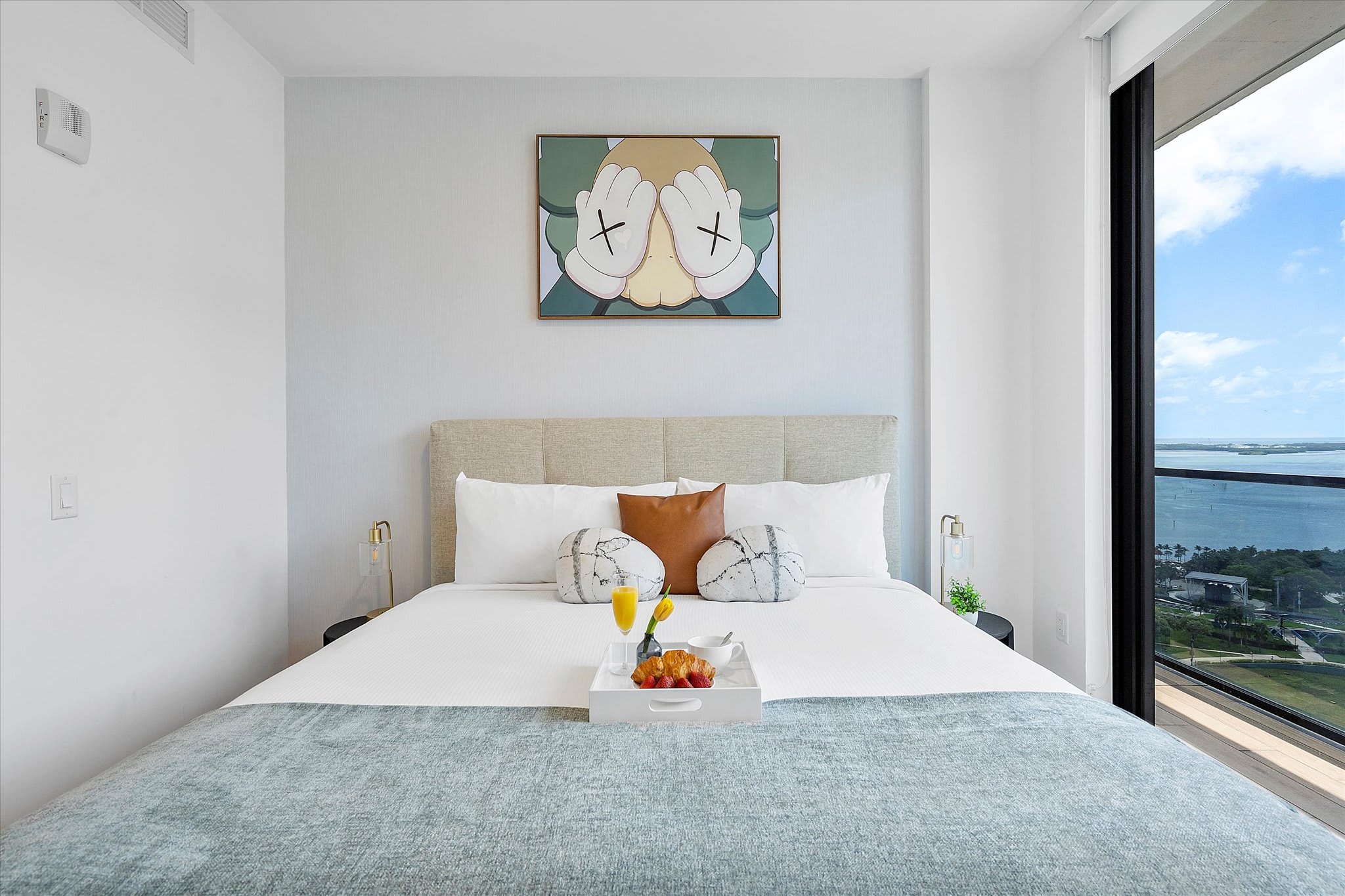 A bright bedroom featuring a king-size bed with a light upholstered headboard, a breakfast tray, and large windows revealing a partial bay view. A distinctive KAWS-style artwork is prominently displayed above the bed.