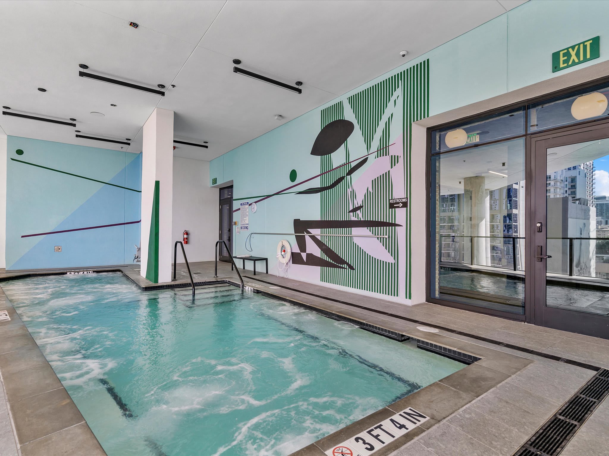 An indoor hot tub area features a large bubbling pool with steps, set against a vibrant mural with abstract green and pink designs. An "EXIT" sign is visible above glass doors leading outside.