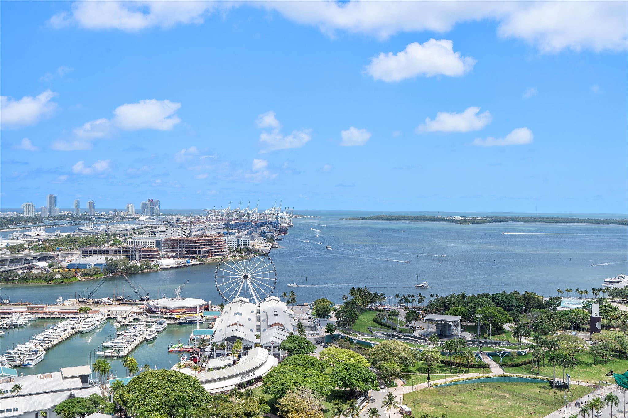 Breathtaking aerial view of a vibrant city bay area, showcasing a marina filled with boats, a large Ferris wheel, and numerous high-rise buildings along the waterfront under a clear blue sky.