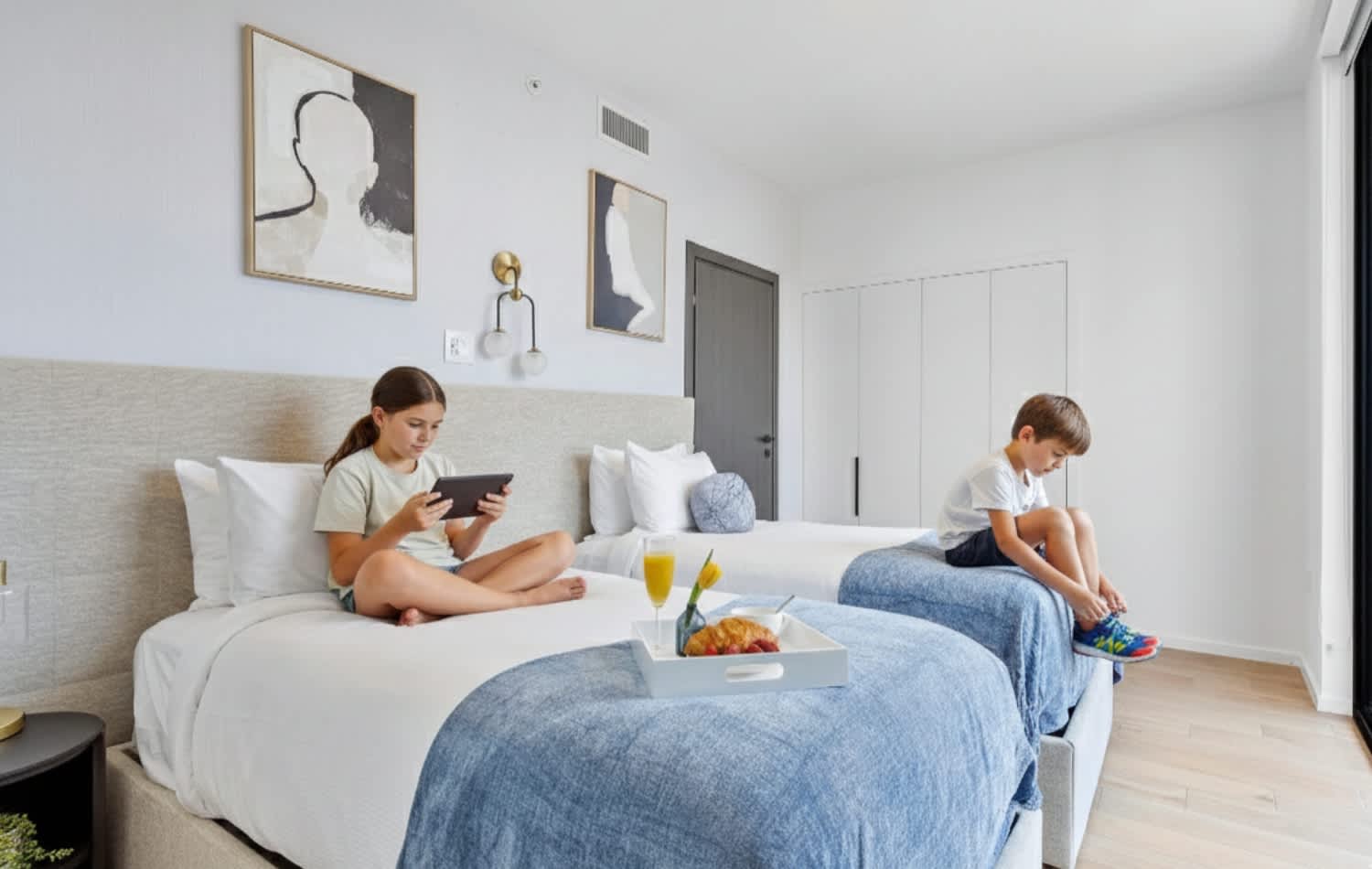 Bright bedroom with two single beds, each with white and blue bedding, and modern artwork on the walls. A girl is on one bed with a tablet, and a boy is on the other, suggesting a family-friendly setup.
