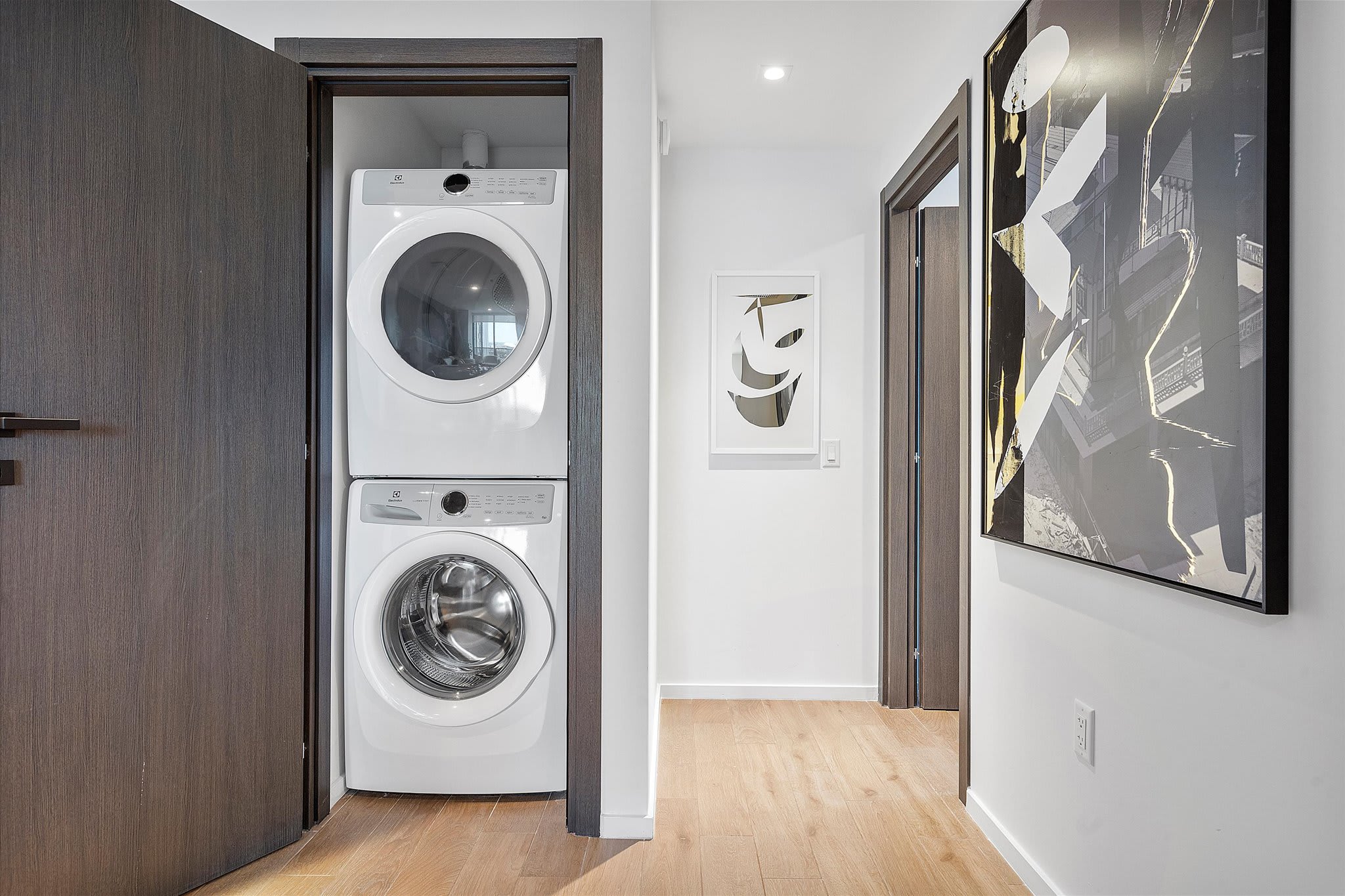 A stacked white washer and dryer unit is neatly tucked into a closet space with dark wood trim. The surrounding hallway features light wood flooring and white walls with modern artwork visible in the distance.