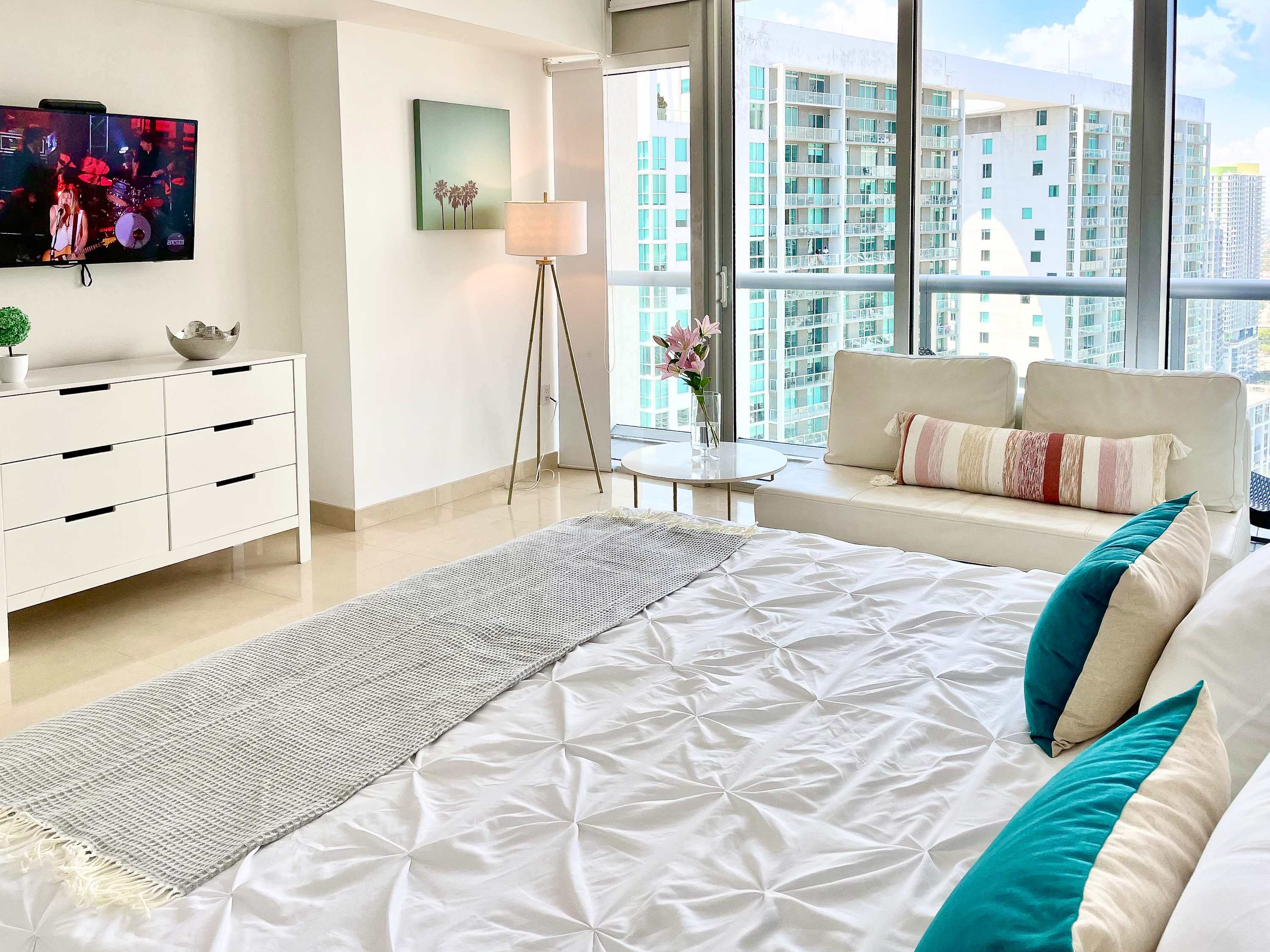 Wide view of the bright bedroom featuring a white queen bed with teal and striped pillows, a white dresser with a TV, and a floor lamp. Floor-to-ceiling windows offer expansive city views.