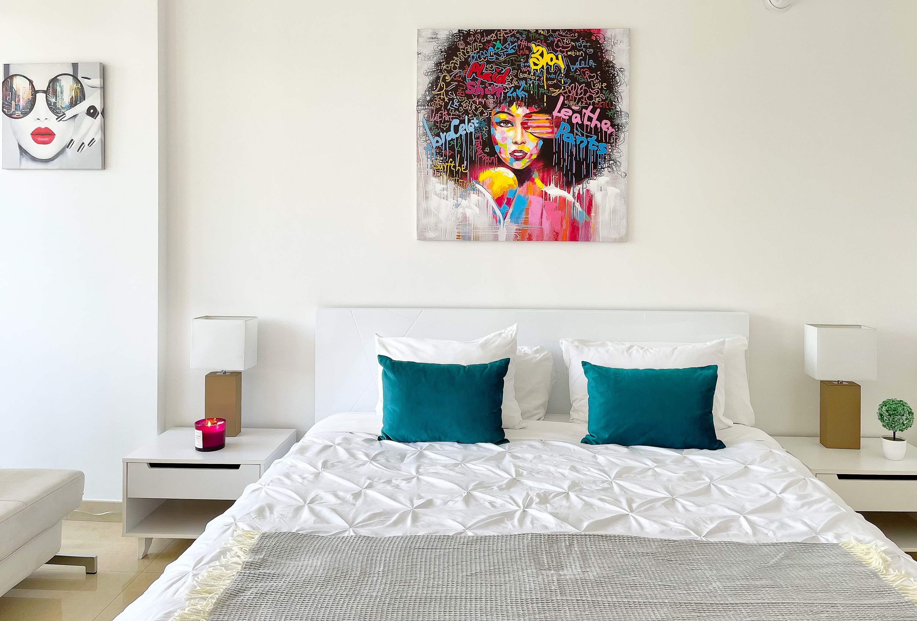 Close-up of the white queen bed with teal throw pillows and a gray blanket, flanked by white nightstands with modern lamps. A vibrant abstract portrait hangs above the headboard, adding a pop of color.