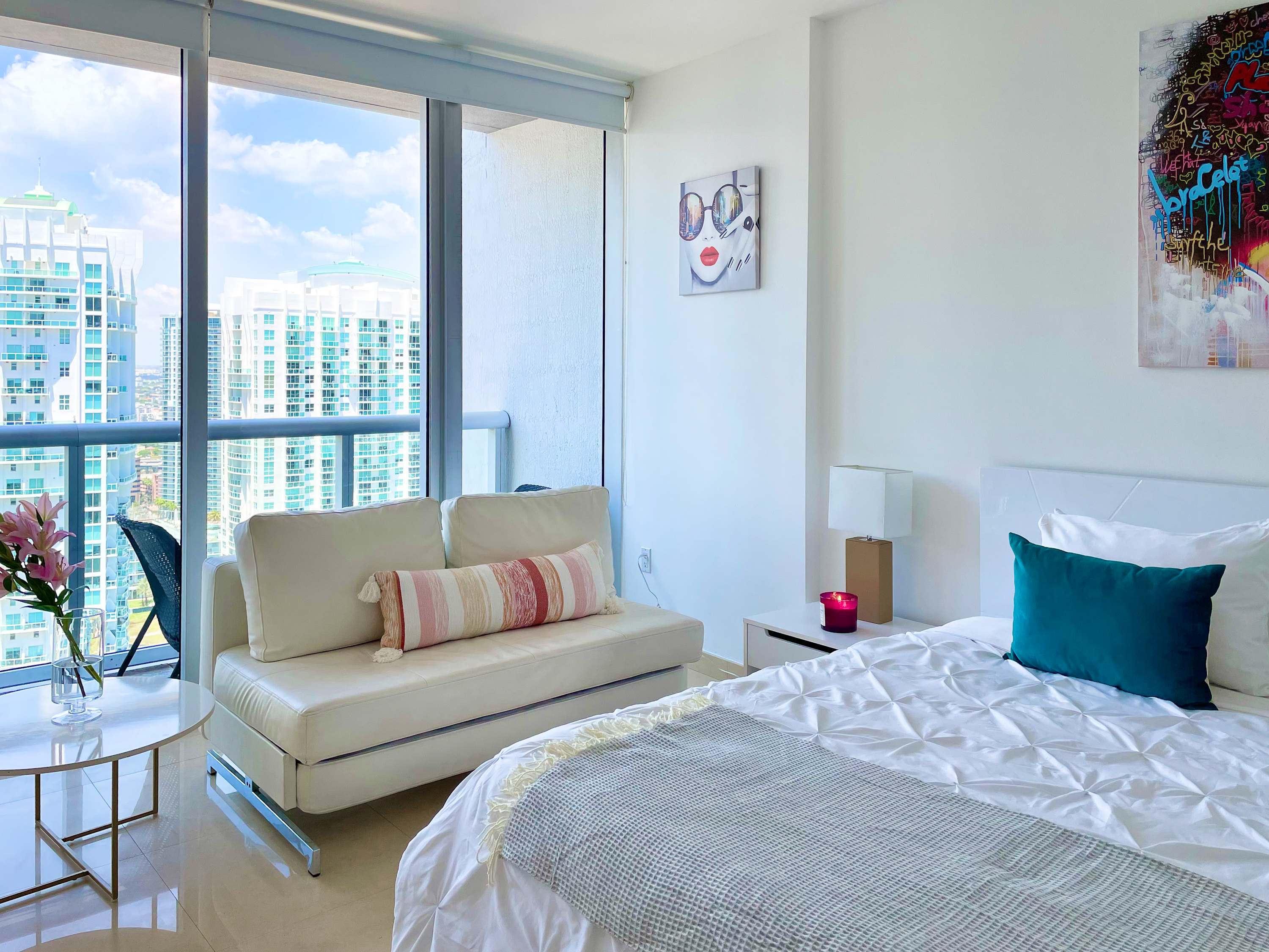 Bright bedroom with a white queen bed, teal pillows, and a gray throw, featuring a vibrant abstract portrait above the bed. A white chaise lounge sits by floor-to-ceiling windows offering city views.