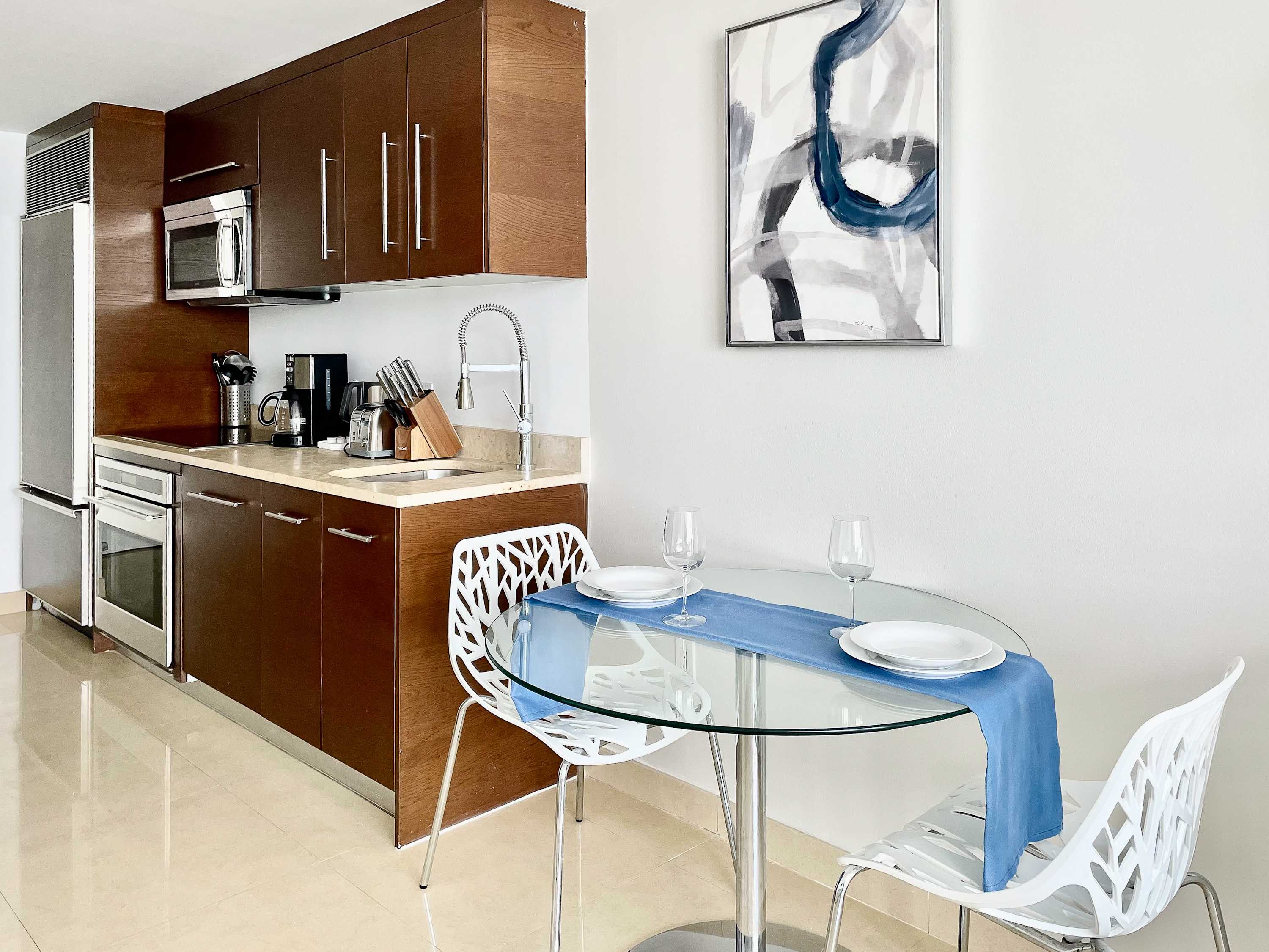 Modern kitchen with dark wood cabinetry, stainless steel appliances, and light-colored countertops. A small glass dining table with two white chairs and a blue runner is set for two, beneath abstract wall art.