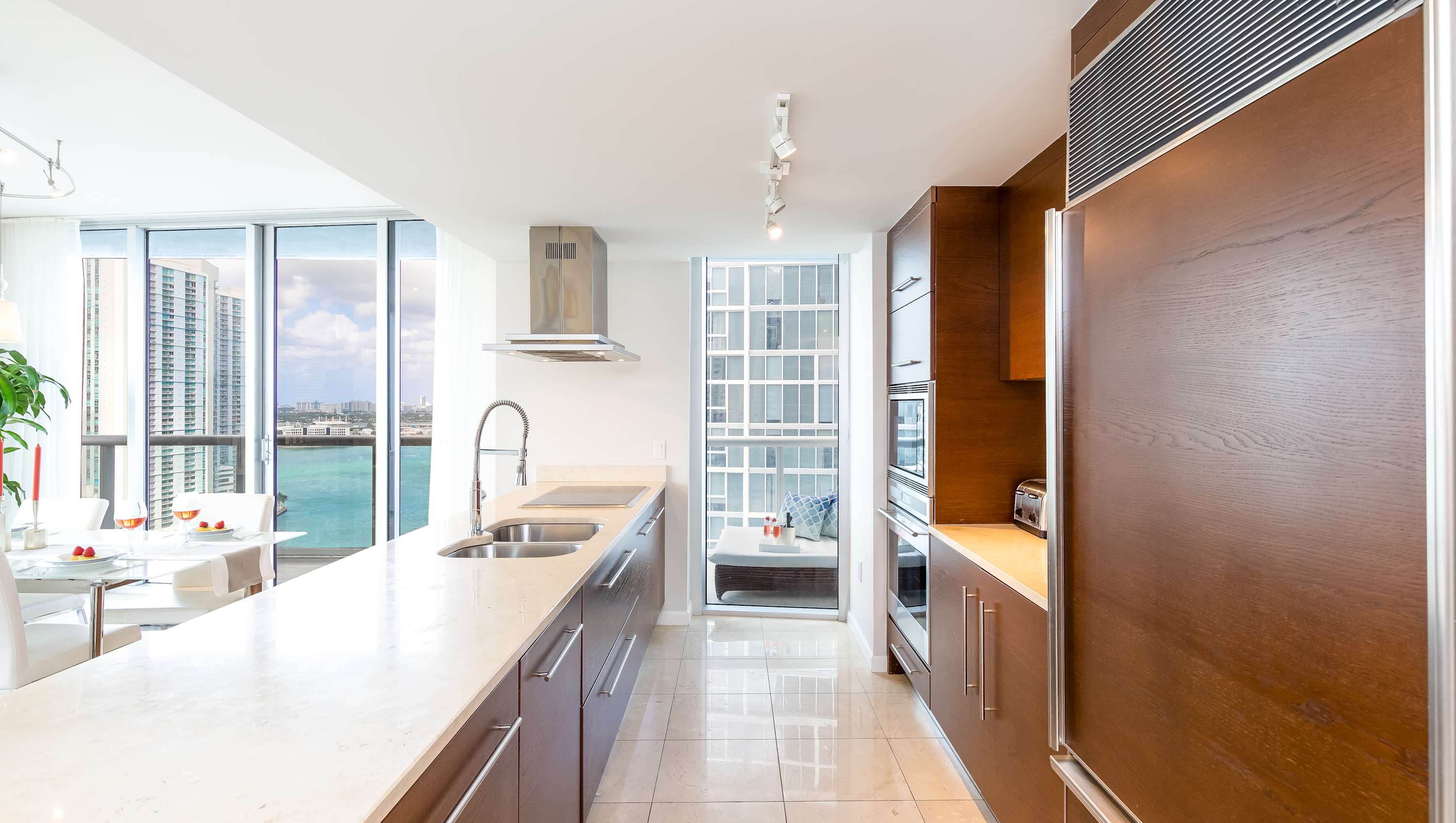Modern kitchen featuring a long island with a light stone countertop, stainless steel sink, and contemporary faucet, complemented by dark wood cabinetry and stainless steel appliances. Floor-to-ceiling windows offer a glimpse of the water view.