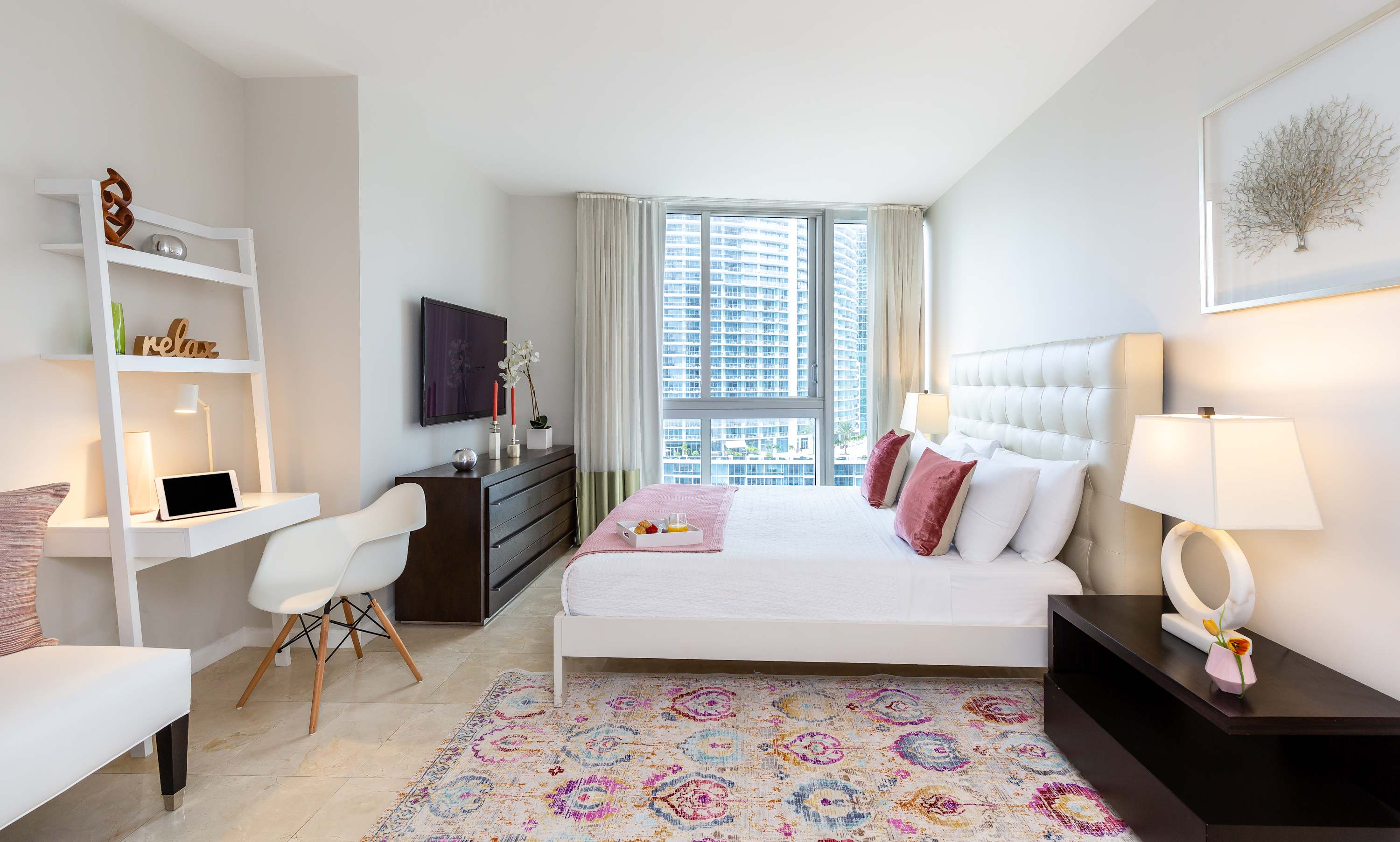 Bright bedroom with a white upholstered queen bed, dark wood dresser, and a white desk area, featuring a colorful patterned rug and large windows with city views. Pink and white accents add a touch of warmth.