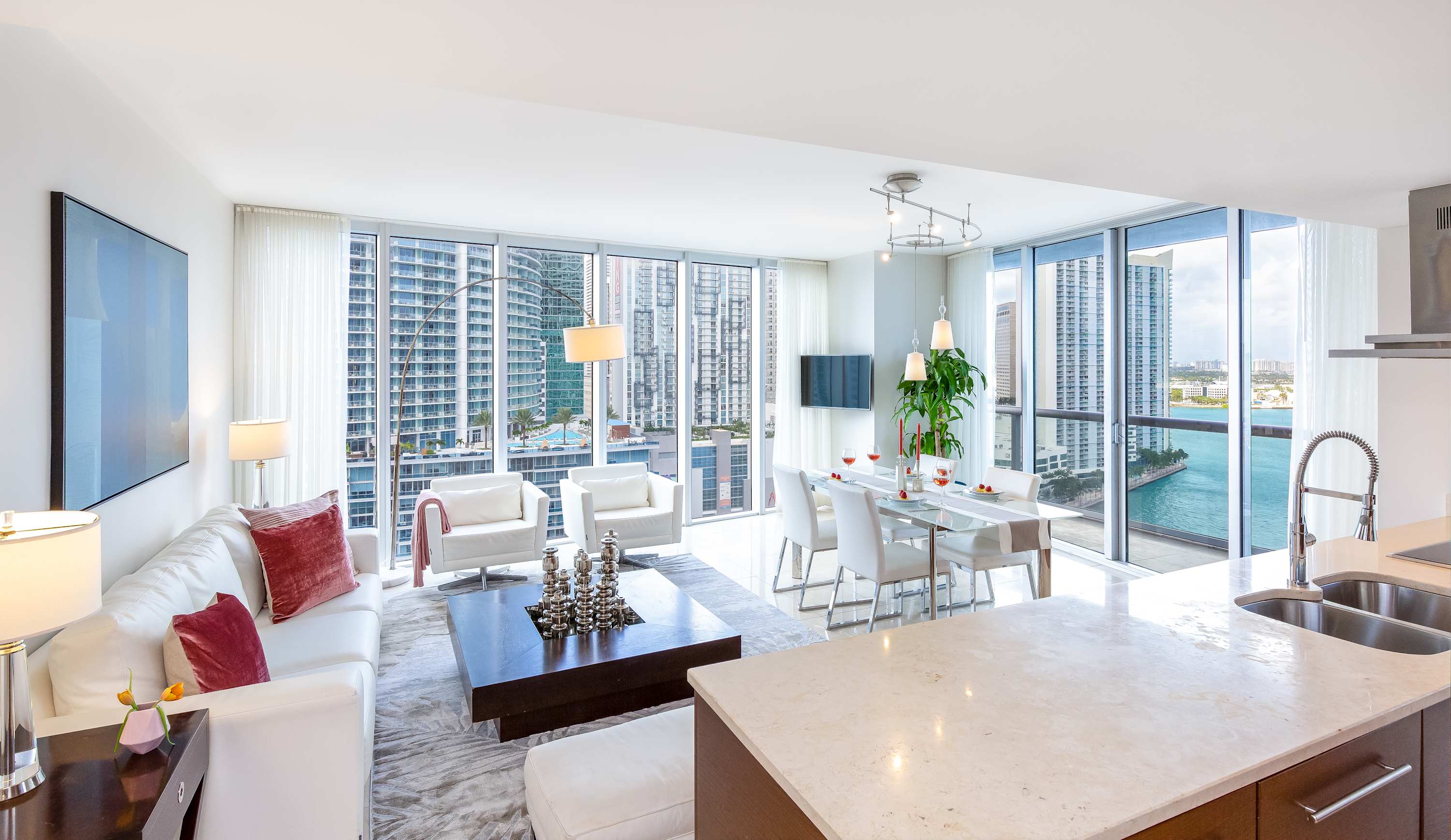 Expansive open-concept living space featuring a white sofa, dark coffee table, and a dining area with a white table, all set against a backdrop of floor-to-ceiling windows with city and water views. A modern kitchen with an island is visible in the foreground.