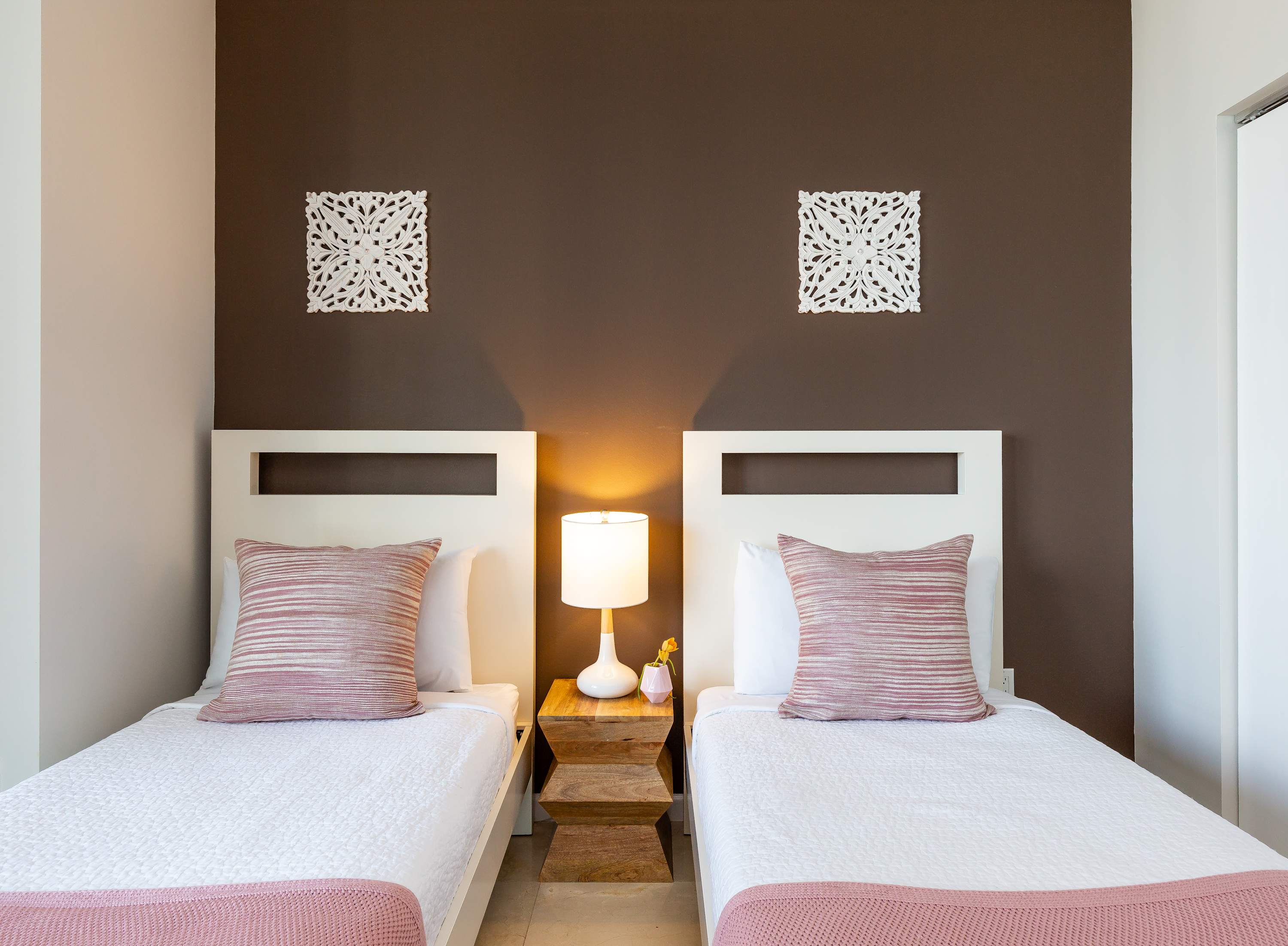 Close-up of two twin beds in a bedroom, each with white bedding, pink throws, and striped pillows, separated by a rustic wooden nightstand with a lamp. A dark brown accent wall is adorned with white decorative panels.