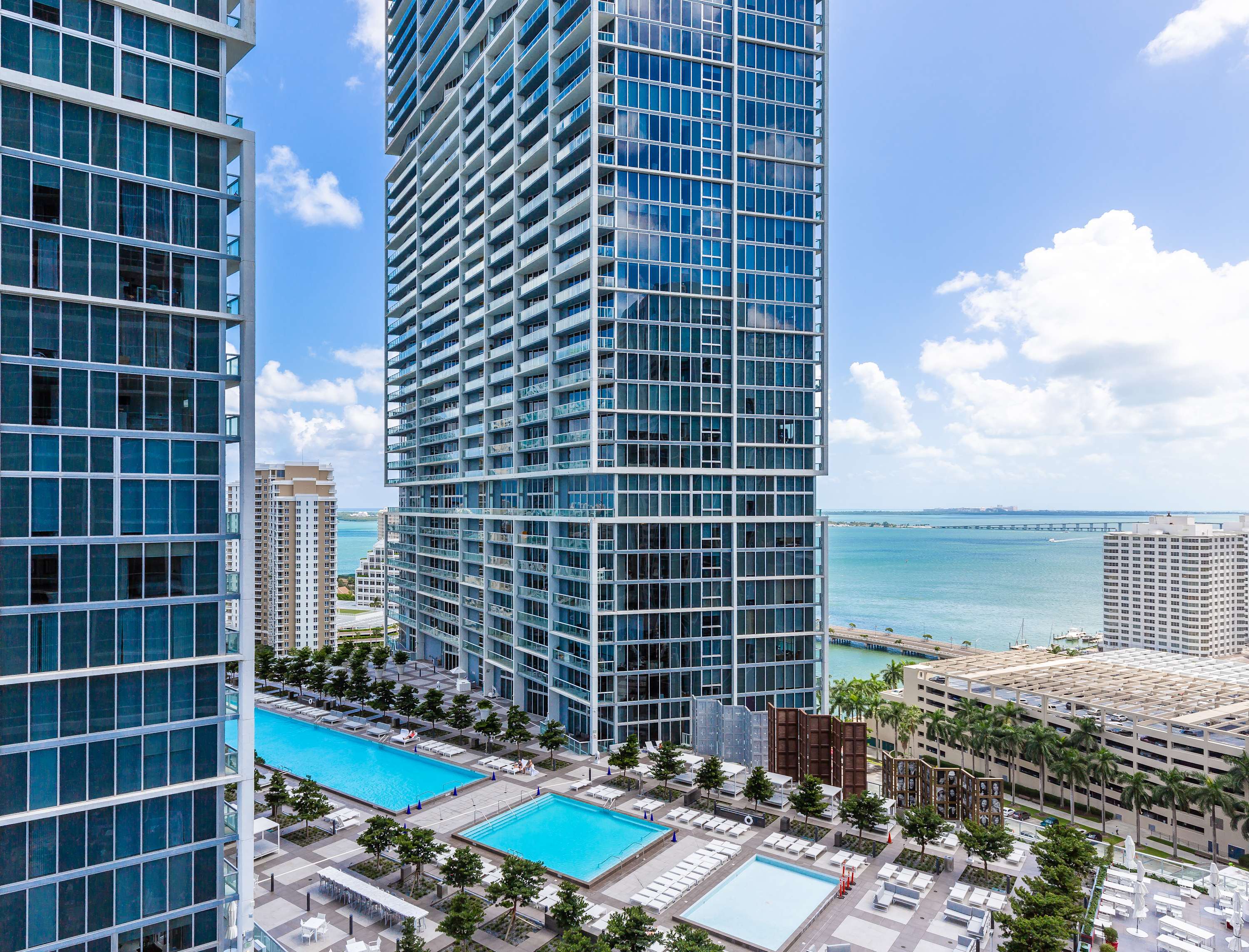 Aerial view of a large outdoor pool deck with multiple swimming pools, surrounded by lounge chairs and palm trees, set against a backdrop of modern high-rise buildings and a bay. The vibrant blue pools contrast with the urban landscape.