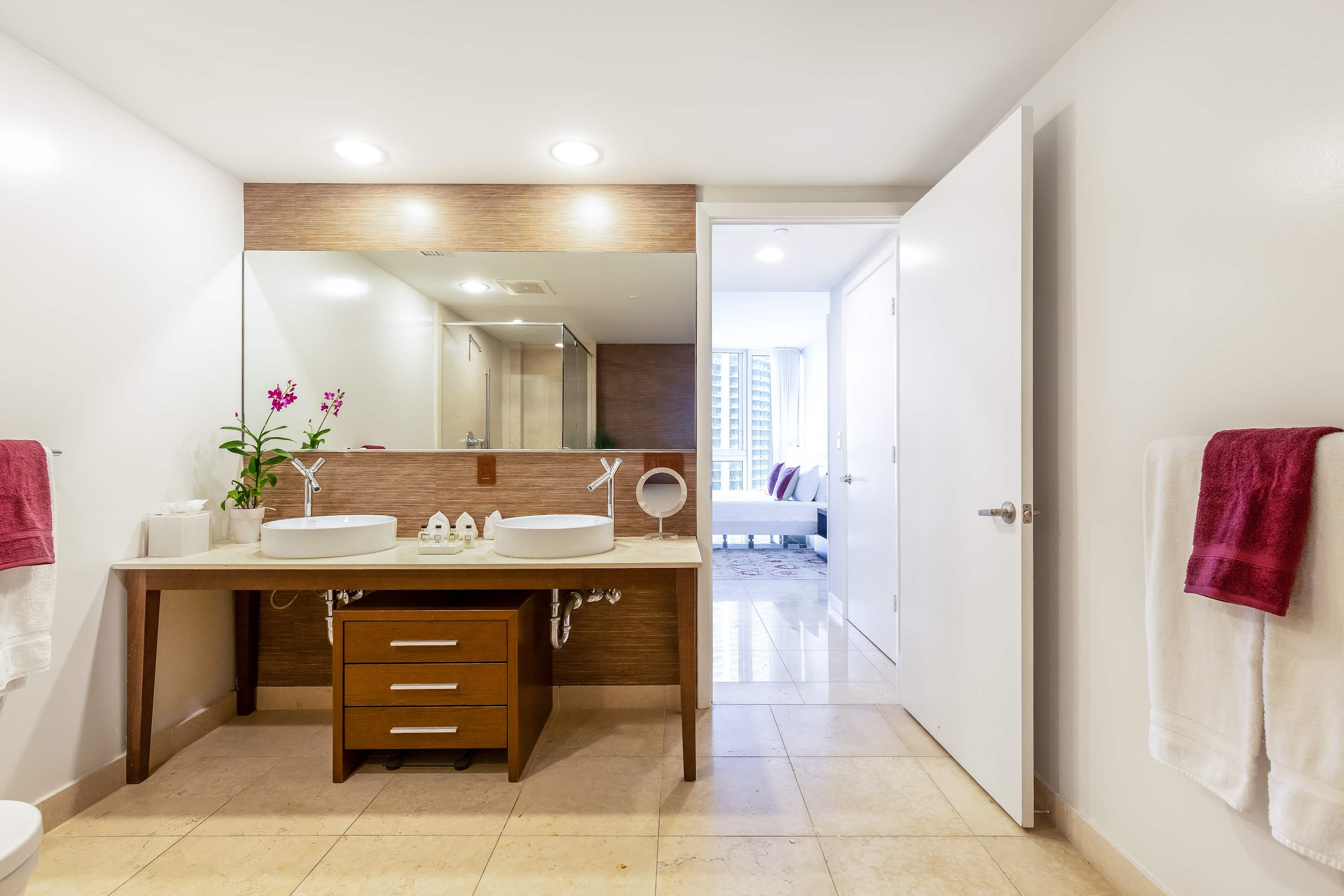 Wide shot of a modern bathroom with a double vanity, white vessel sinks, and a large mirror, leading into a bright bedroom through an open doorway. The vanity features a dark wood base with drawers, and the walls are a mix of light paint and horizontal brown tiling.