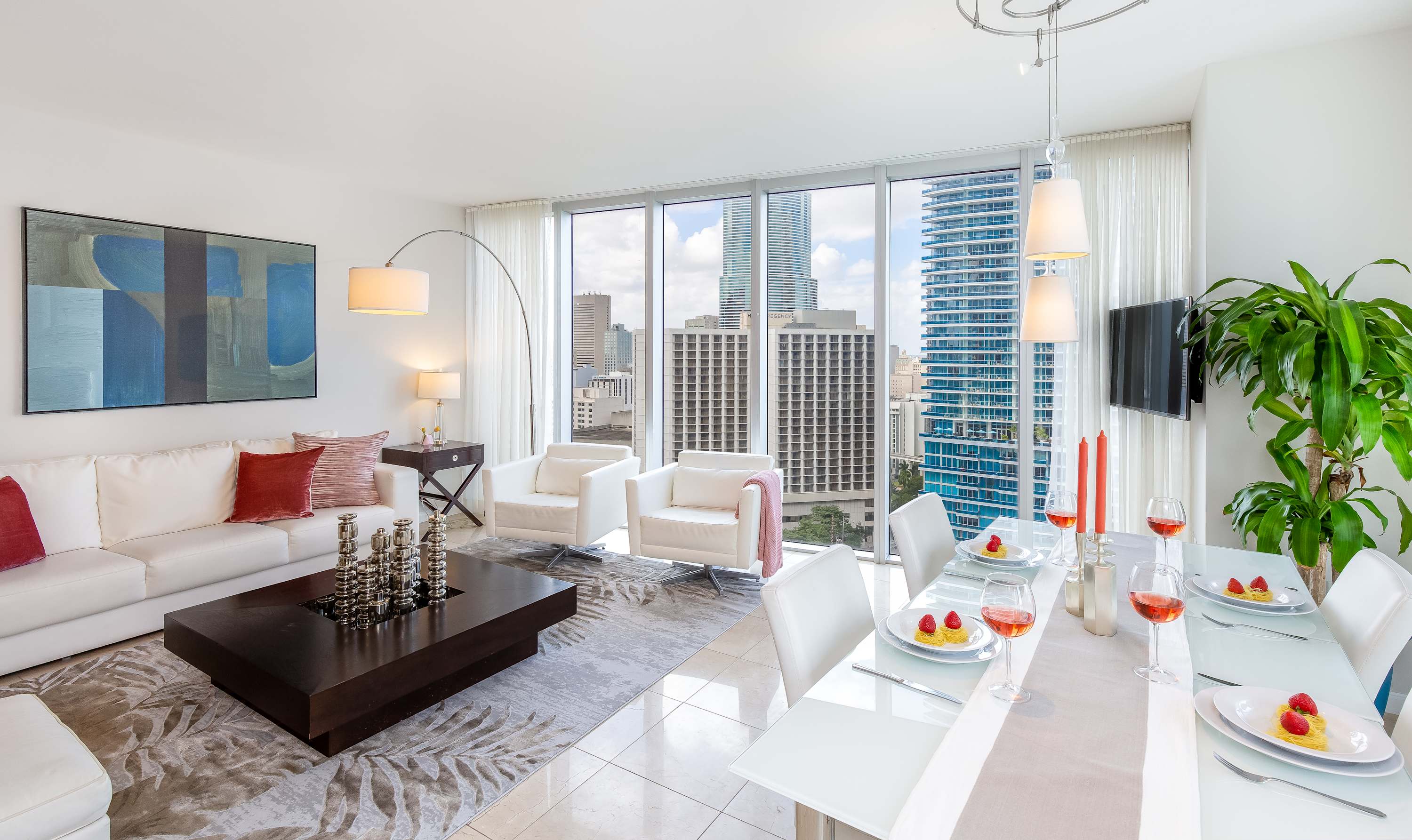 Bright, open-concept living and dining area with white furniture, large windows offering city views, and a modern aesthetic. The living space features a white sofa, dark coffee table, and a large abstract painting.