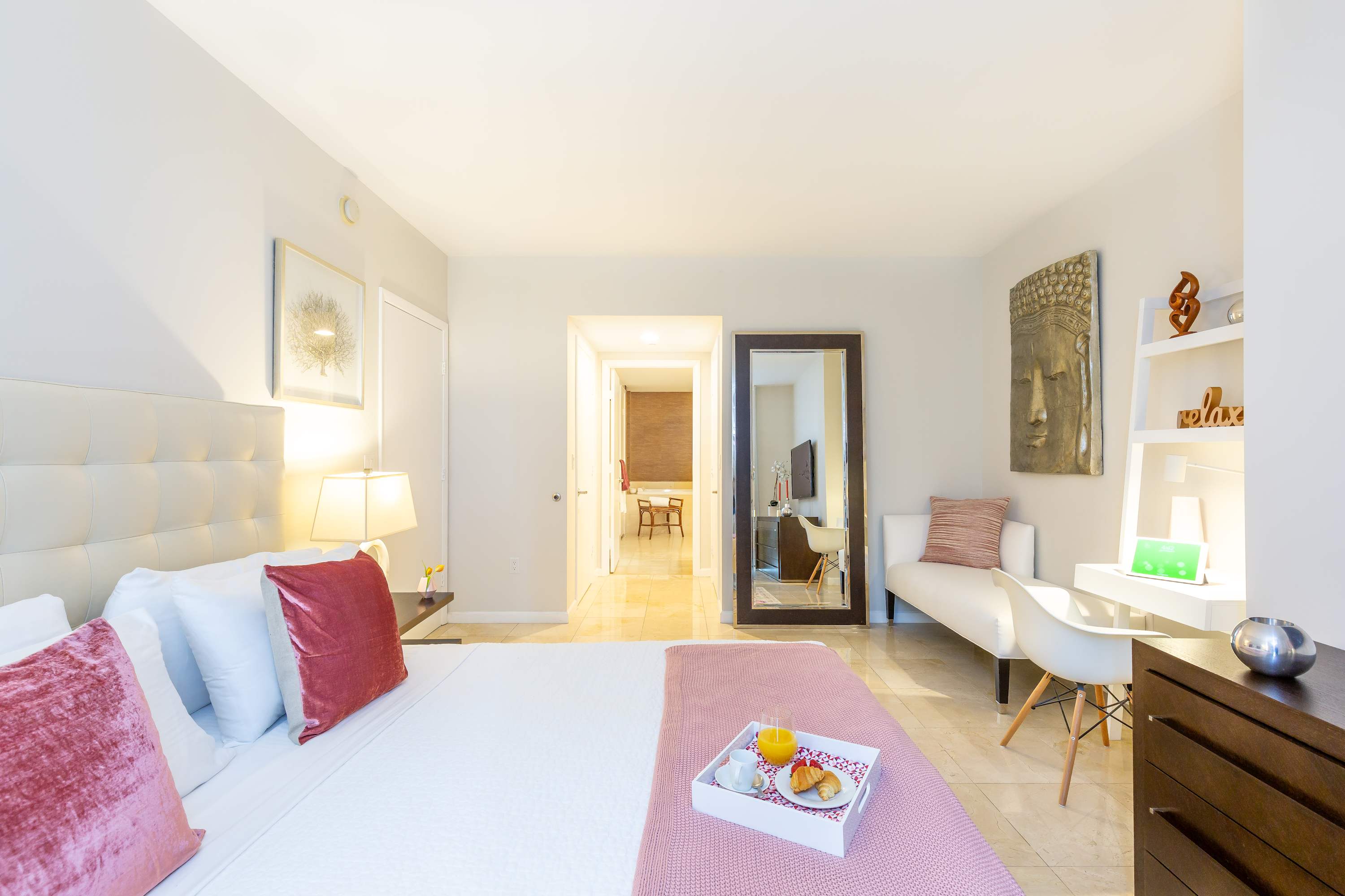 Master bedroom with a white upholstered queen bed, a white desk and shelving unit, and a full-length mirror, featuring light tile floors and a colorful area rug. A large Buddha head sculpture adds an artistic touch.