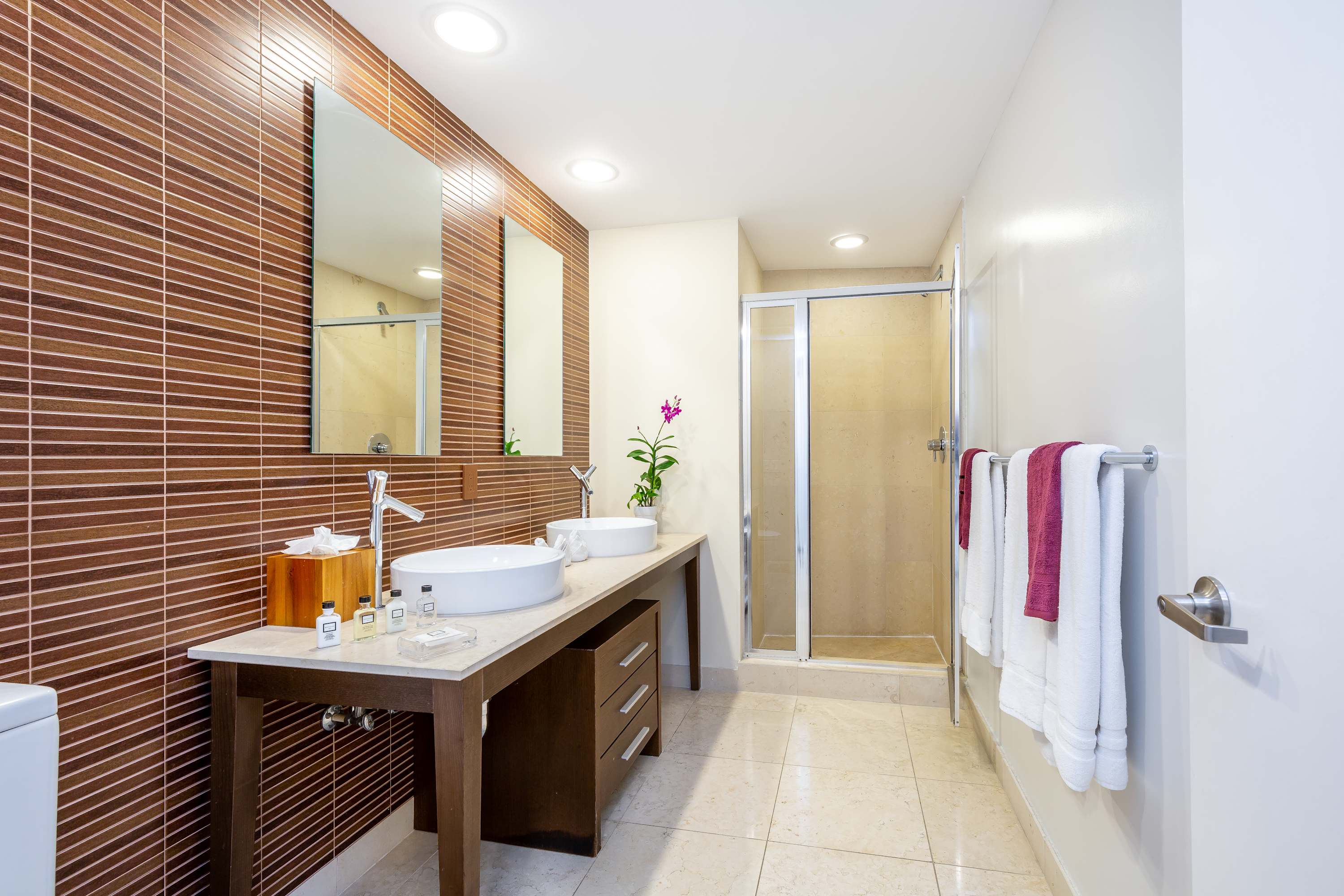 Modern bathroom featuring a double vanity with white vessel sinks, a large mirror, and a walk-in shower with a glass enclosure. The wall behind the vanity is tiled with horizontal brown strips, contrasting with the light-colored floor tiles.