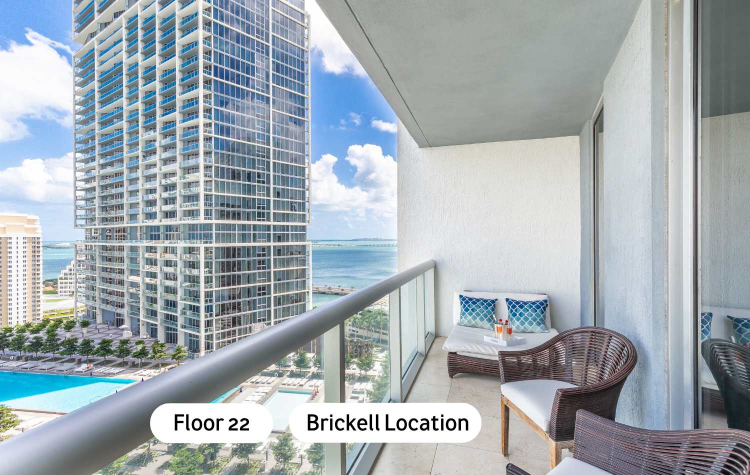 A modern balcony with glass railings offers panoramic city and bay views, featuring a wicker lounge chair and a small table with drinks. A large high-rise building and a pool area are visible below.