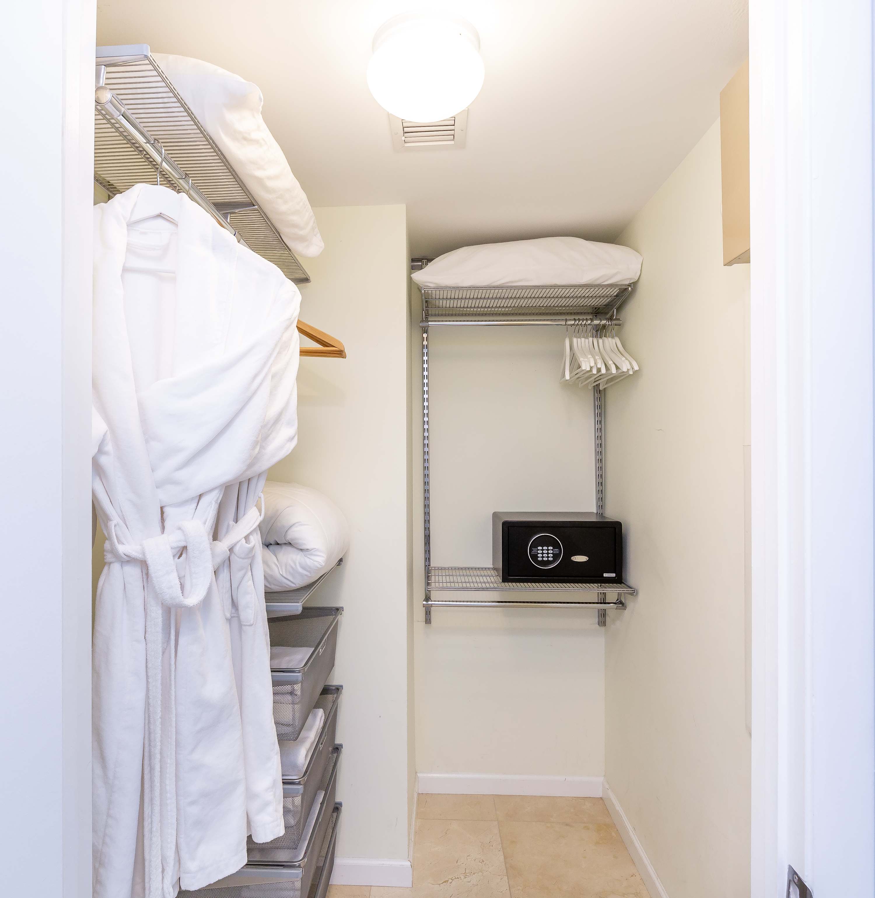 Walk-in closet with wire shelving units, a white bathrobe hanging, and a small black safe. Extra pillows and blankets are stored on the shelves, and the floor is tiled.