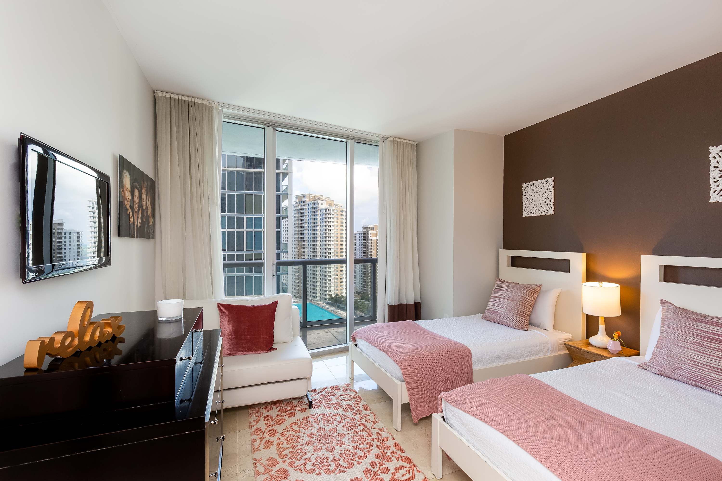 Second bedroom featuring two white twin beds with pink throws and striped pillows, a dark brown accent wall, and large windows offering city and pool views. A dark dresser with a TV and a white lounge chair complete the space.