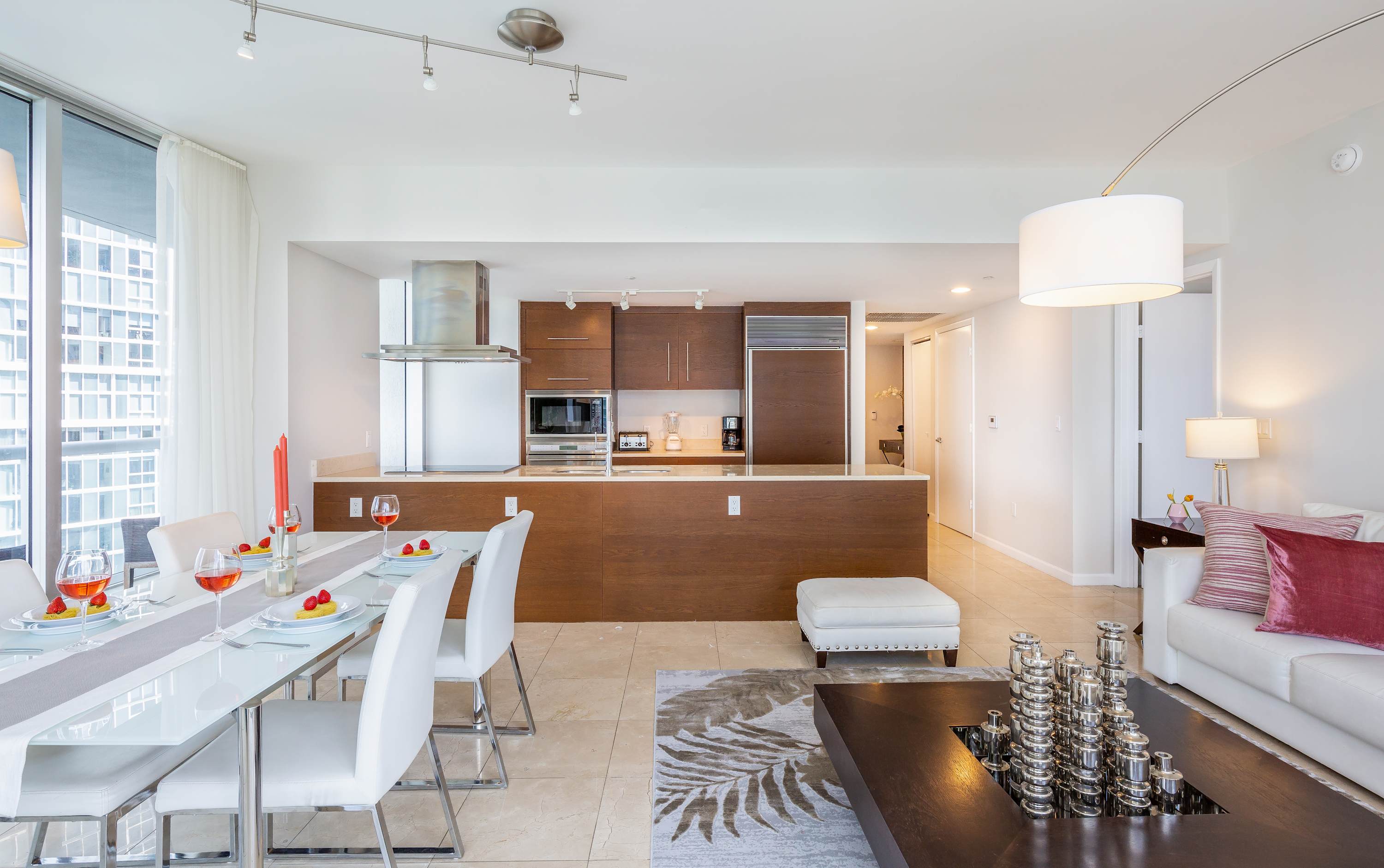Modern open-plan kitchen with dark wood cabinetry, stainless steel appliances, and a light-colored countertop, seamlessly flowing into a dining area with a white table set for four. The space features light tile flooring and contemporary lighting.