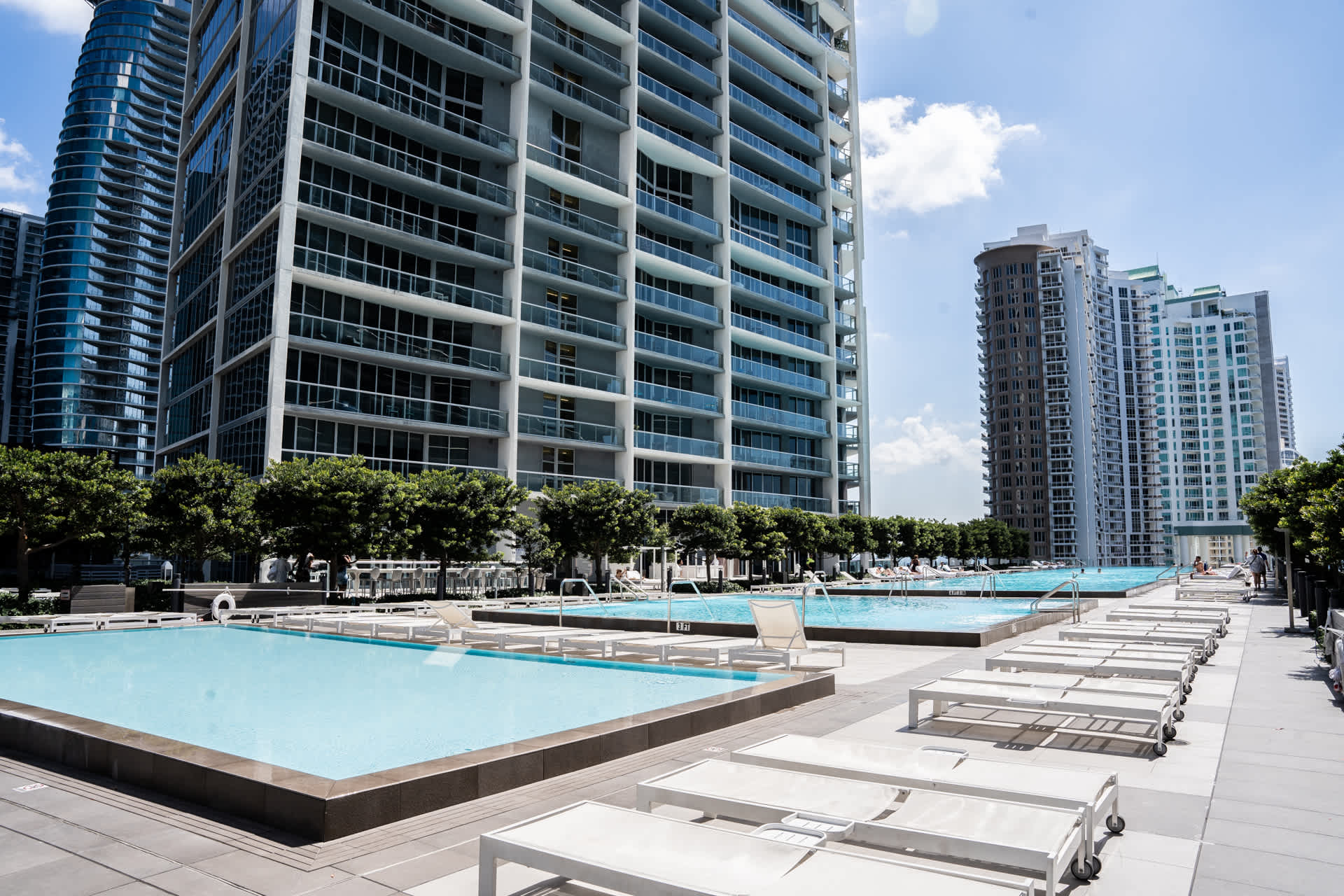 Expansive outdoor pool deck featuring multiple rectangular swimming pools with clear blue water, surrounded by numerous white lounge chairs. Tall modern high-rise buildings frame the sunny urban landscape.