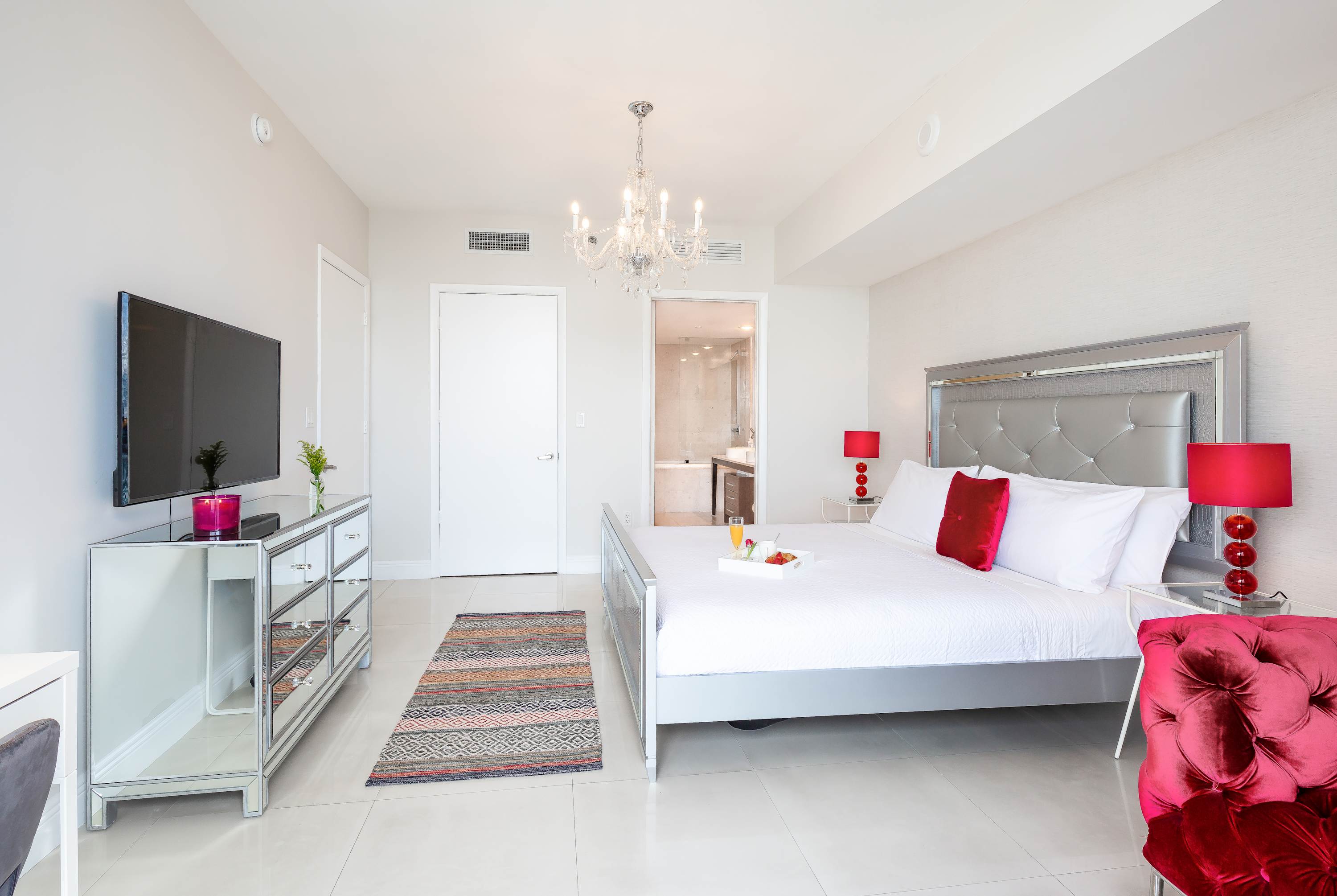 Wide shot of bedroom-1 showcasing the bed with a breakfast tray, a mirrored dresser with a TV, and a vibrant red tufted armchair. A doorway leads to the bathroom, and a colorful patterned rug accents the white tiled floor.