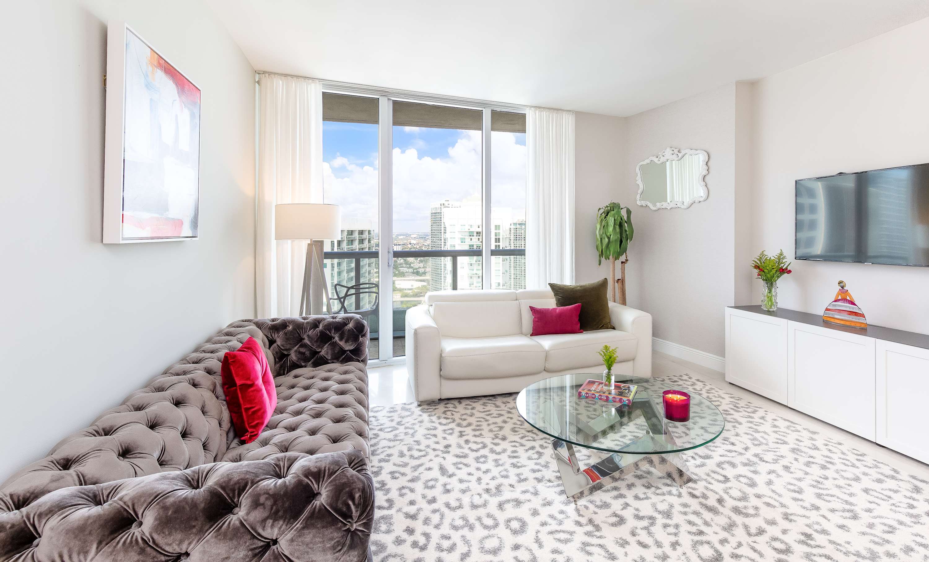 Bright living room featuring a grey tufted velvet sofa and a white modern sofa, a glass coffee table, and a large sliding glass door leading to a balcony with city views. A patterned rug covers the light-colored tiled floor.