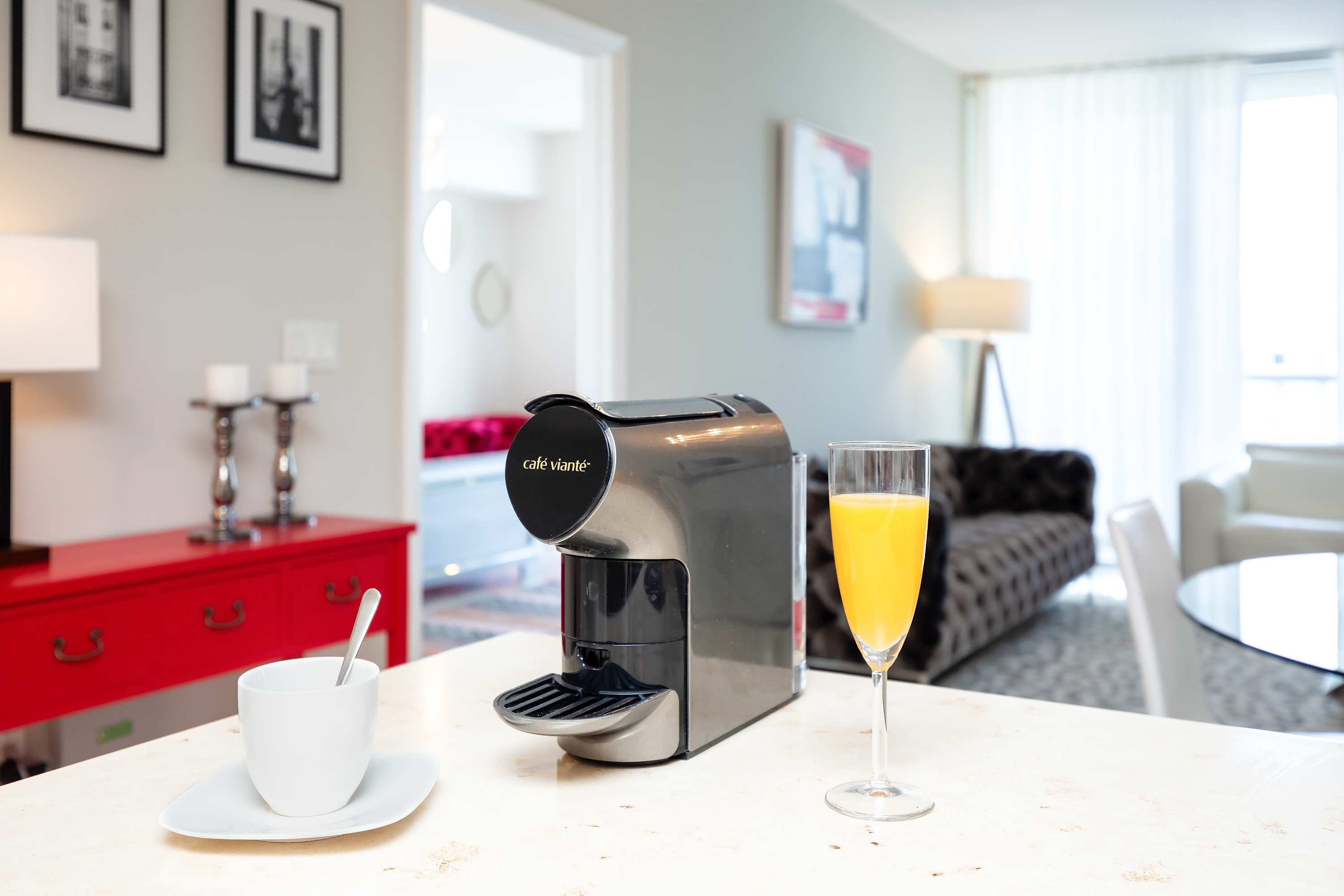 A coffee machine and a glass of orange juice sit on a white countertop, with a glimpse of the living area featuring a dark tufted sofa and a dining table in the background.