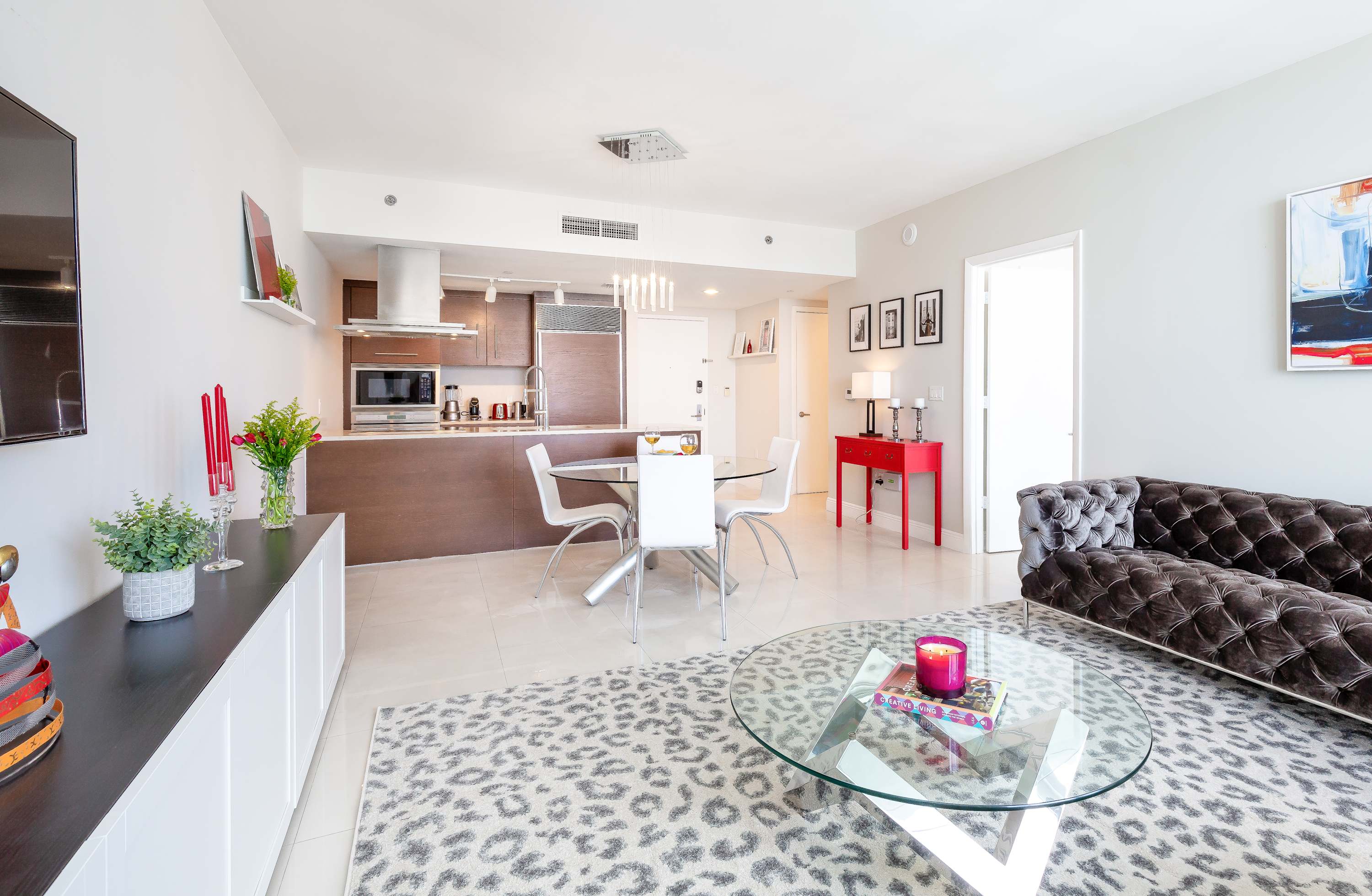 View of the open-concept living space from the dining area, showing a dark tufted sofa, a glass coffee table, and an area rug. The kitchen with dark wood cabinets is visible in the background.