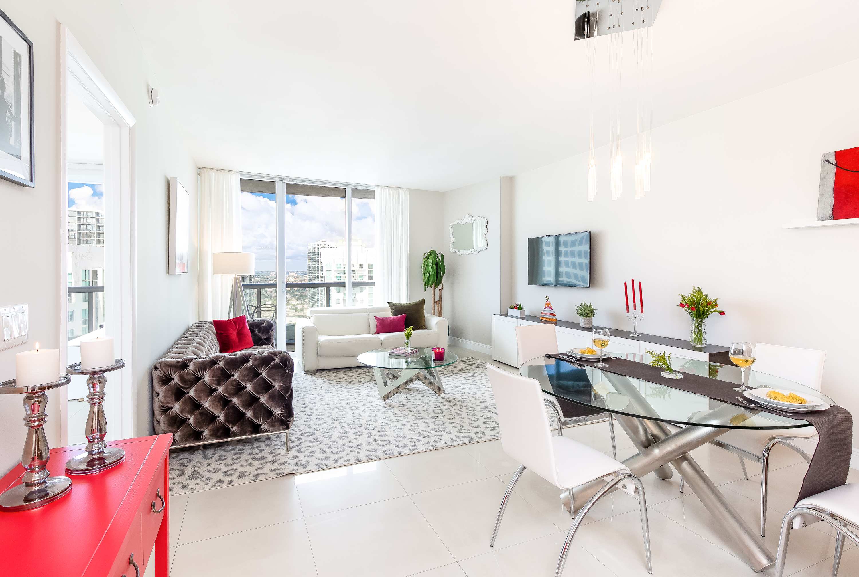 Open-concept living and dining area with a white sofa, dark tufted sofa, glass coffee table, and a glass dining table with white chairs. Large windows offer city views.