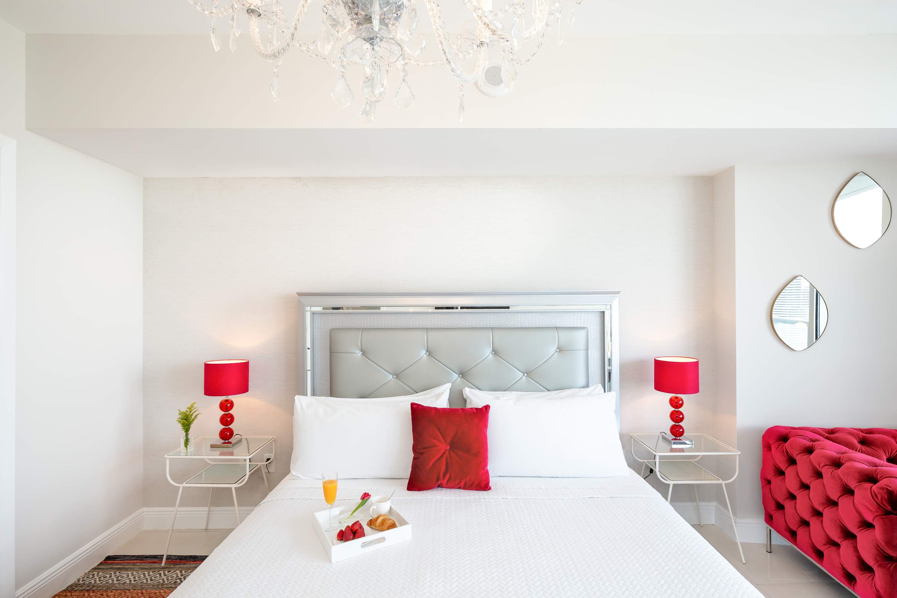 Close-up view of the bed in bedroom-1, featuring a grey upholstered headboard, crisp white bedding with a red accent pillow, and two glass-top nightstands with red lamps. A white tray with breakfast items is placed on the bed.