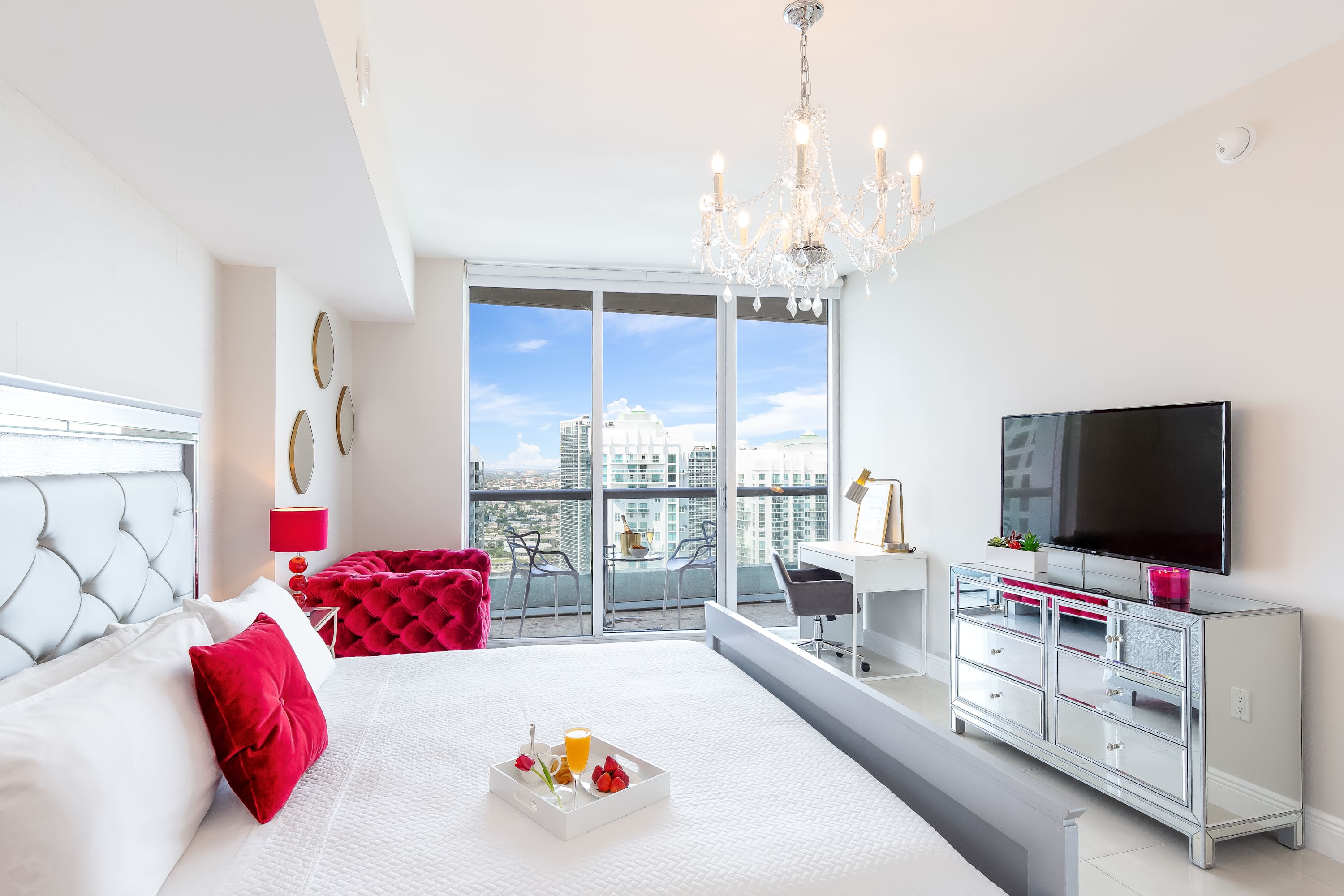 Modern bedroom with a large bed, a mirrored dresser with a TV, a red tufted armchair, and a desk by the window. A large glass sliding door opens to a balcony offering expansive city views.