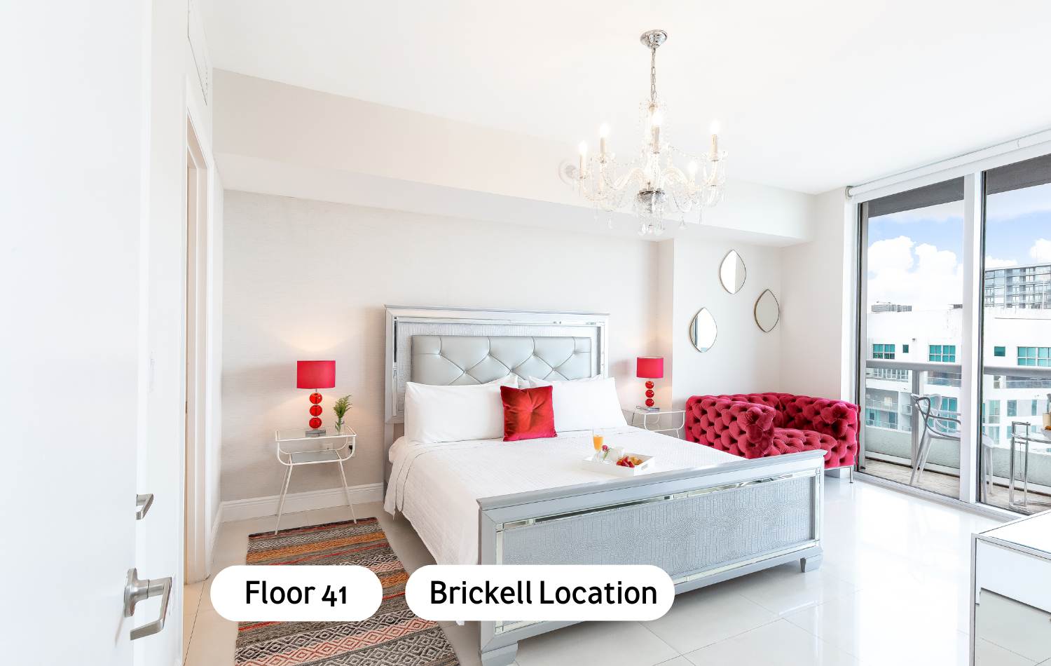 Bright bedroom with a silver-framed bed, white bedding, and red accents from lamps and a throw pillow. A red tufted sofa sits by a large window offering city views.