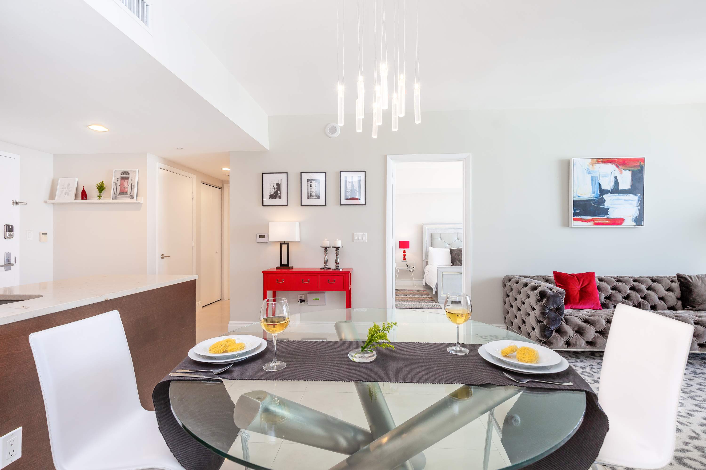 Modern dining area with a round glass table and white chairs, set with two place settings and wine glasses. A red console table and artwork are visible in the background, leading to a bedroom.