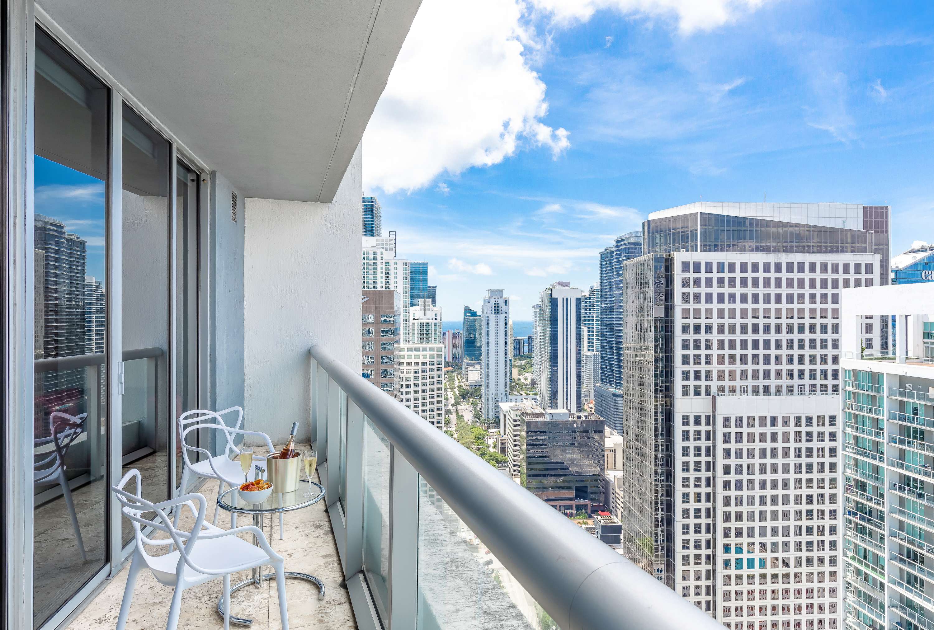 High-rise balcony with a small glass-top table and two white chairs, offering expansive city views of multiple skyscrapers under a partly cloudy sky.