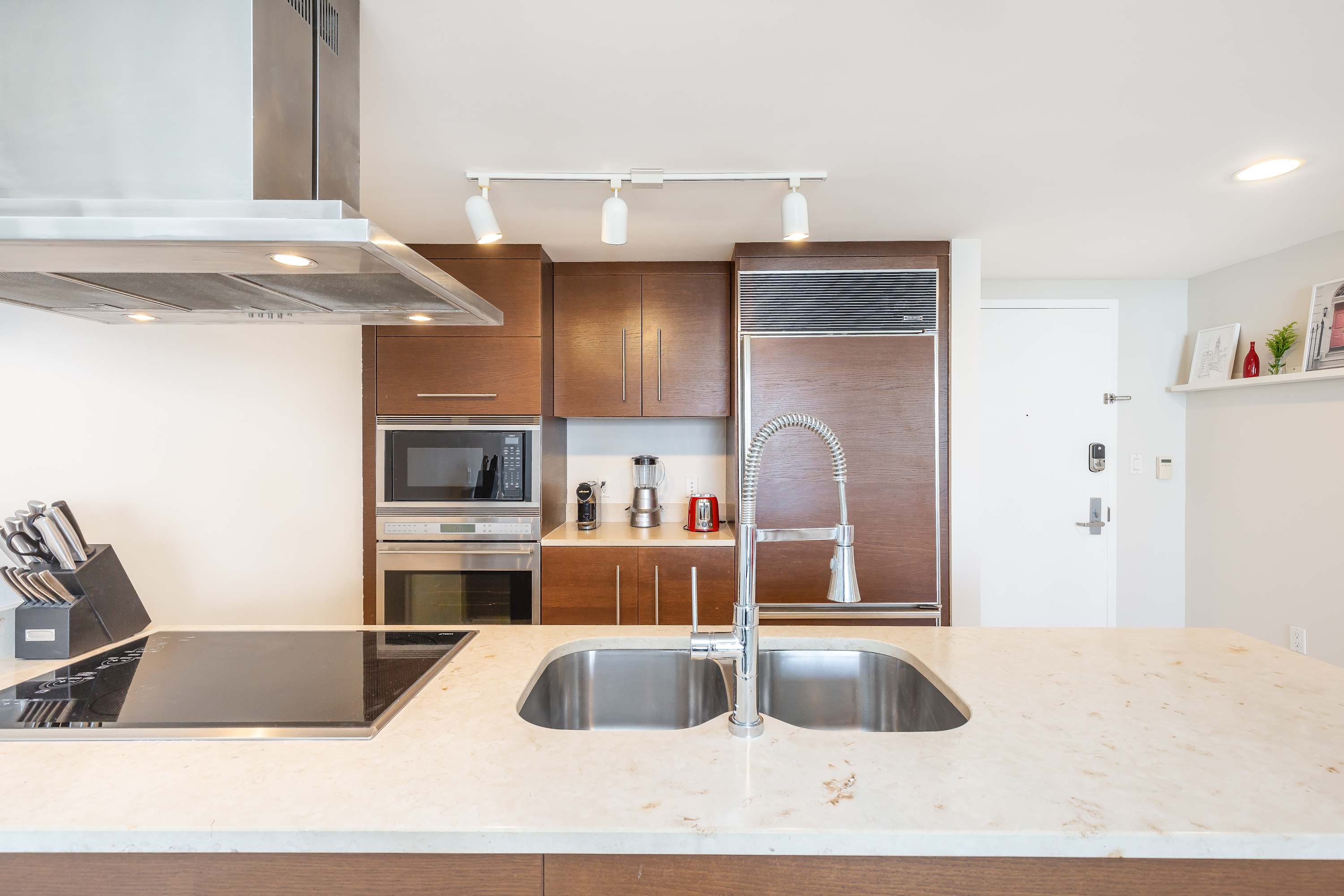 Modern kitchen featuring dark wood cabinetry, stainless steel appliances, and a light marble countertop with a double sink and gooseneck faucet. An induction cooktop and range hood are prominent.