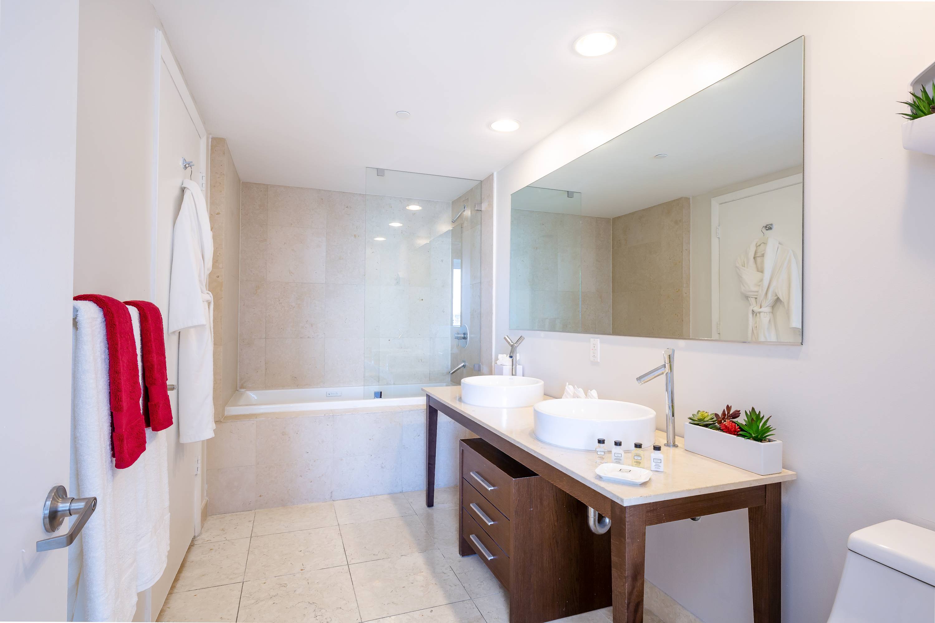 Spacious modern bathroom featuring a double vanity with two white vessel sinks, a large mirror, and a built-in bathtub with a shower. The walls and floor are tiled in a light, neutral stone-like material.