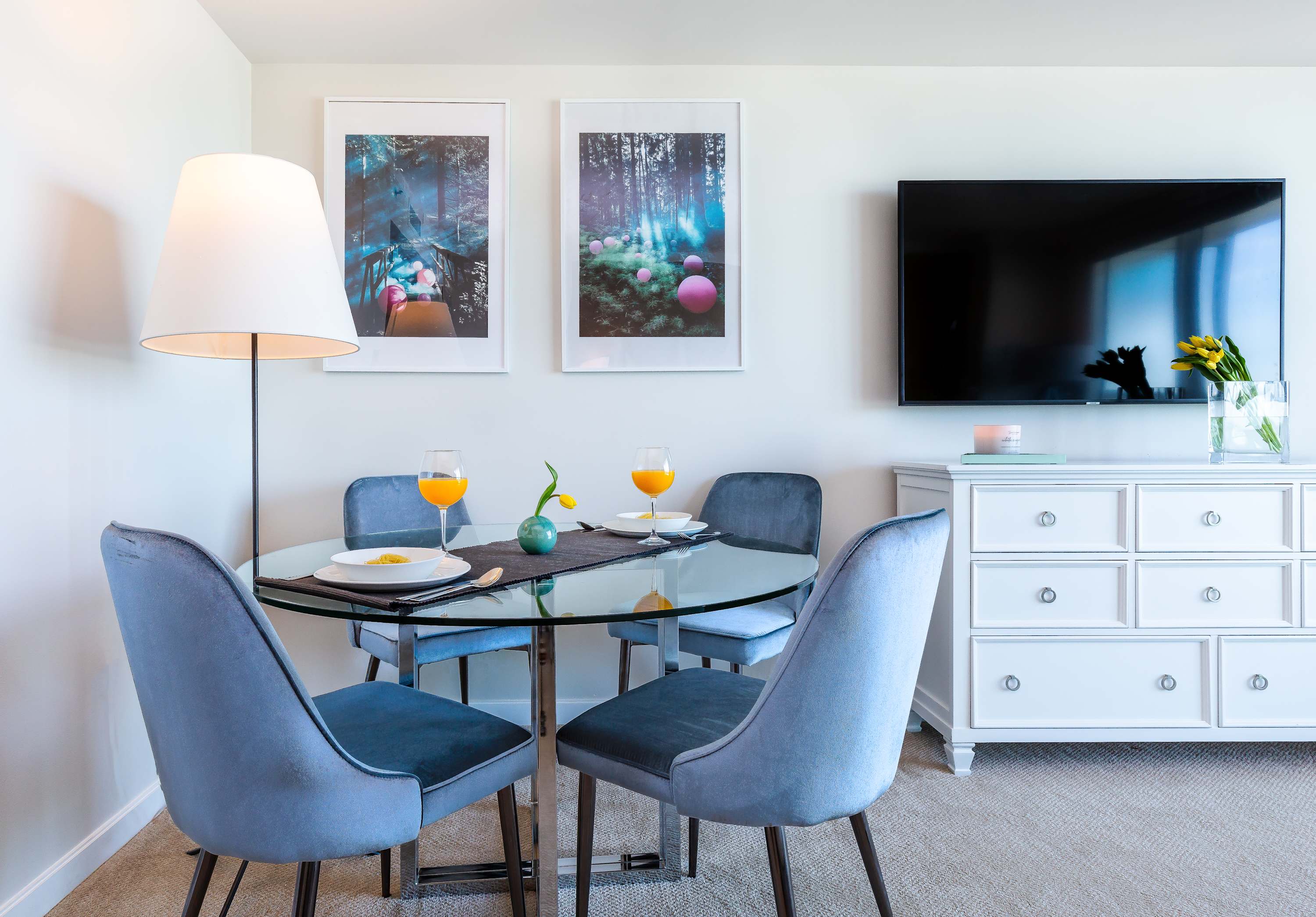 A cozy dining area featuring a round glass-top table with a chrome base and four blue velvet chairs. Two framed art prints hang above a white dresser, and a flat-screen TV is mounted on the wall nearby.