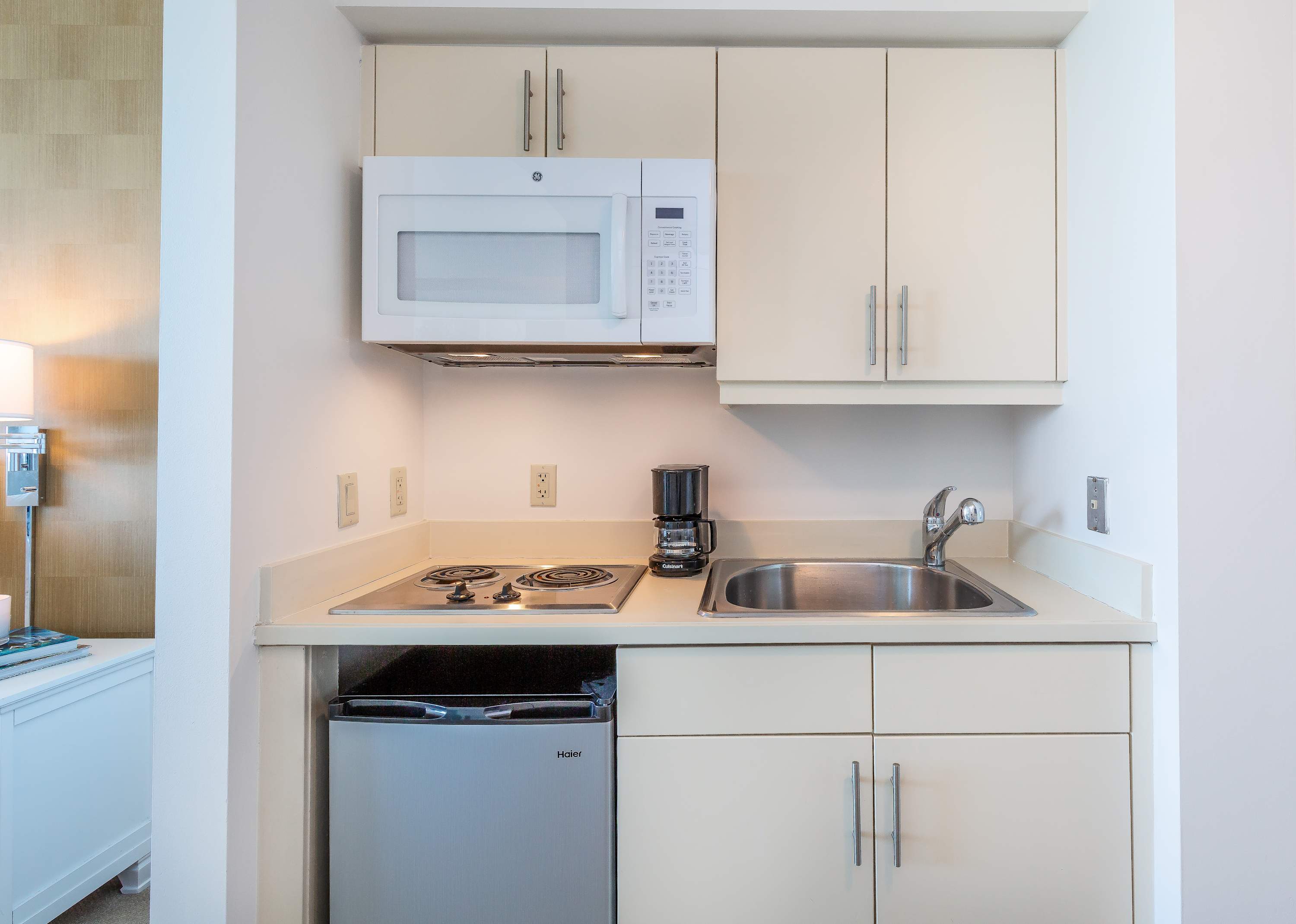 A compact, modern kitchenette with light beige cabinetry, a white microwave, a two-burner electric stovetop, a stainless steel sink, and a small refrigerator. A coffee maker is also visible on the counter.