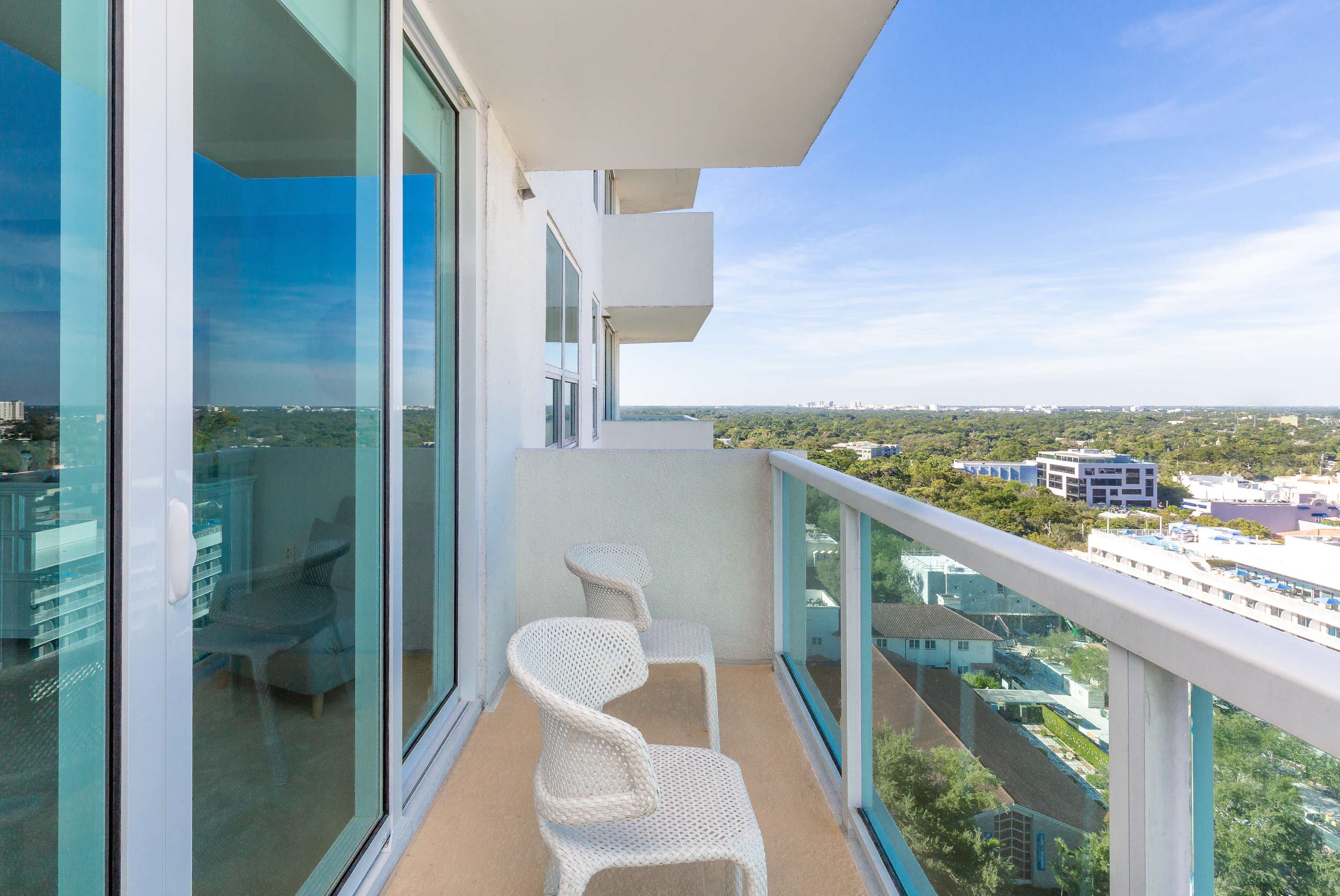 A private balcony with white wicker chairs, offering a panoramic city view with distant buildings and green landscapes under a bright blue sky. Large glass sliding doors connect the balcony to the interior space.