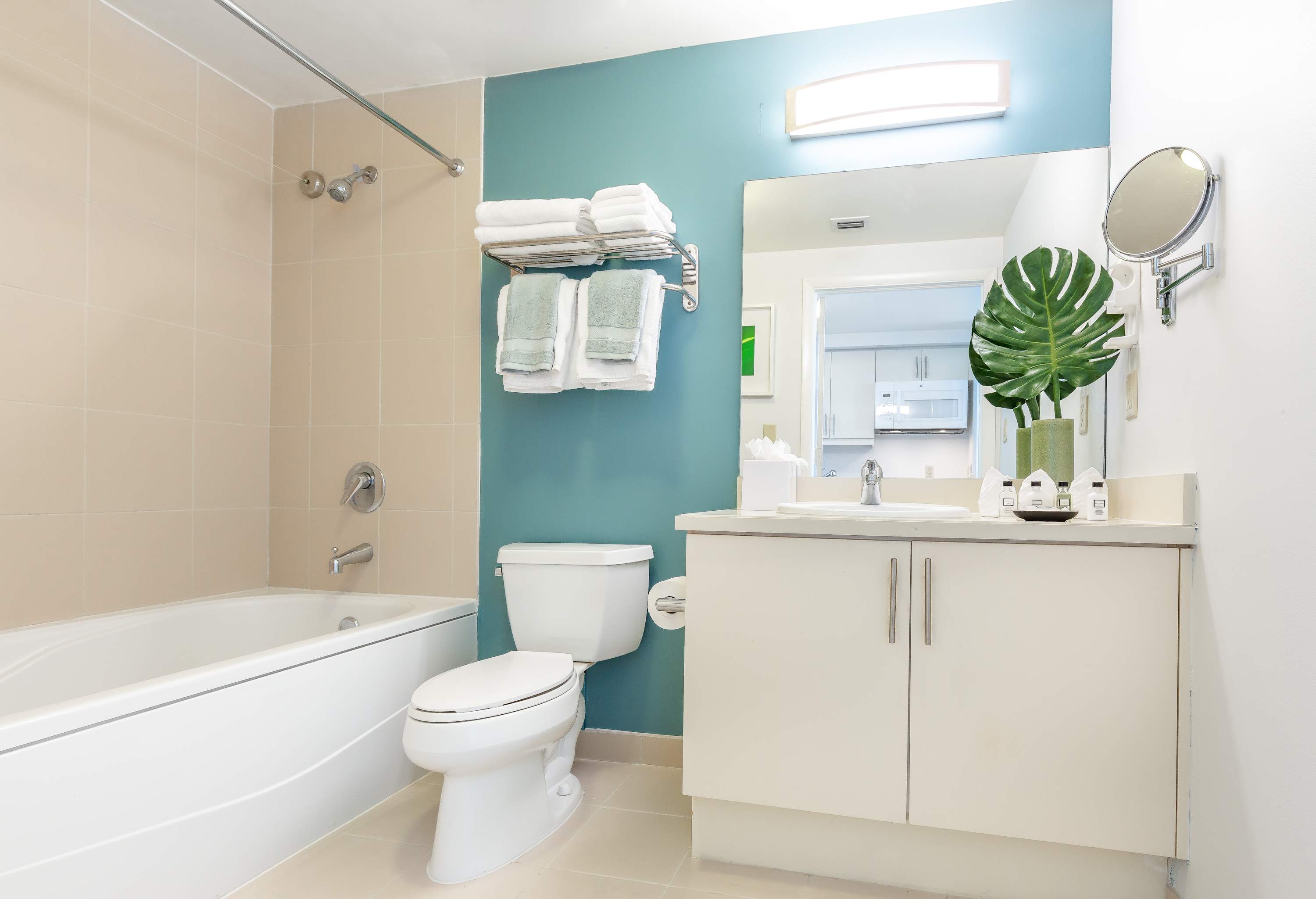 Modern bathroom featuring a white bathtub with a shower, a toilet, and a vanity with a large mirror, set against beige tiled walls and a teal accent wall.