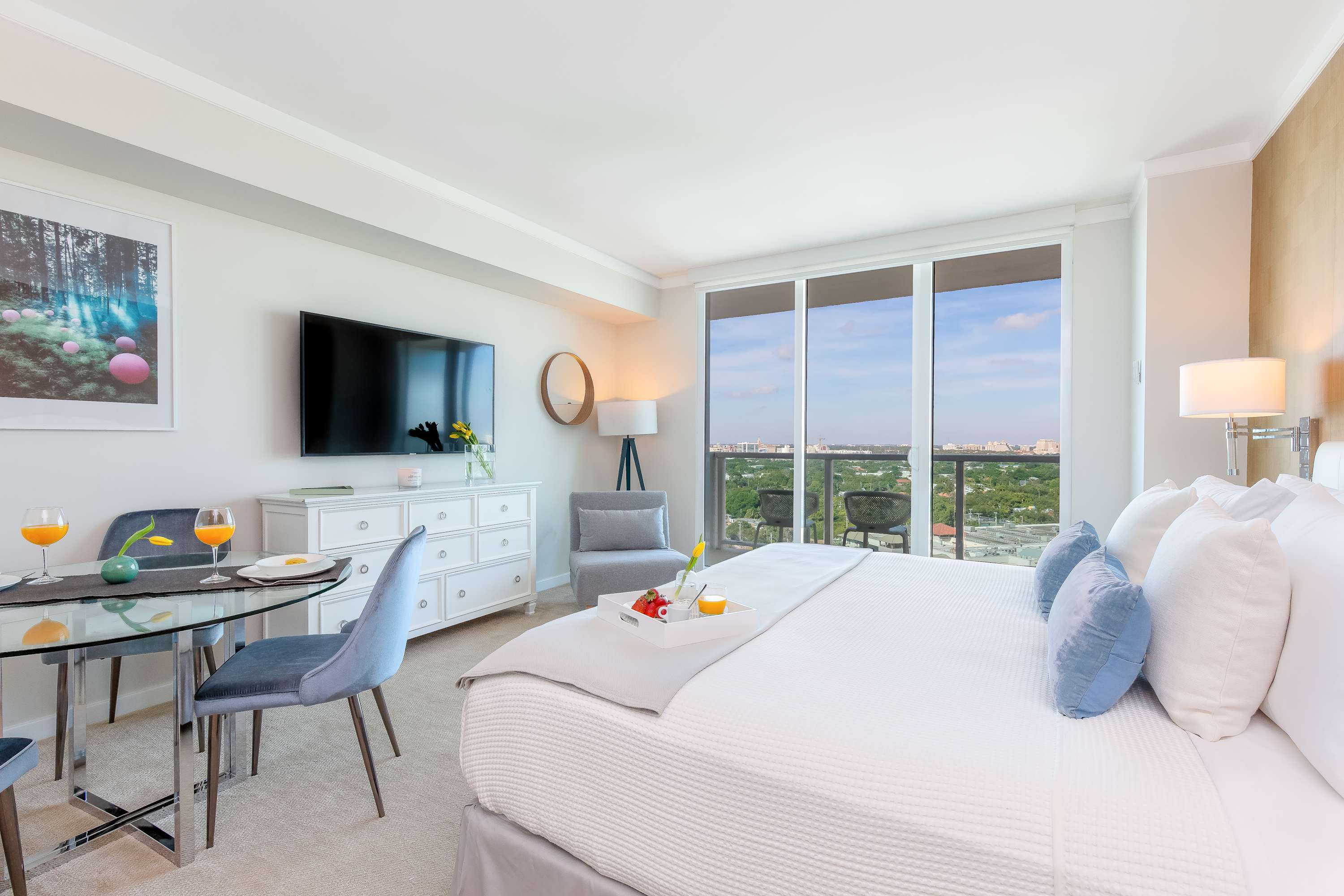 A bright studio apartment showcasing a king-sized bed with white and blue bedding, a glass dining table with blue chairs, and a wall-mounted flat-screen TV. A large window provides a city view, and the room is carpeted with light-colored walls.