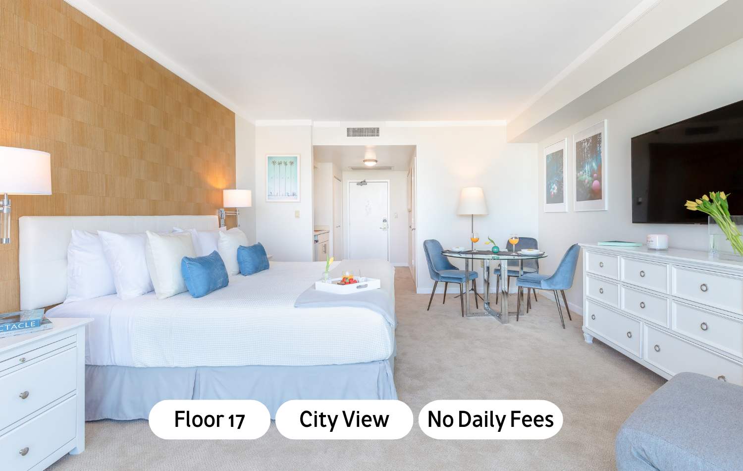 A bright studio apartment featuring a king-sized bed with white and blue bedding, a small dining area with a glass-top table and blue chairs, and a kitchenette visible in the background. The room has light-colored walls, carpeted floor, and a textured accent wall behind the bed.