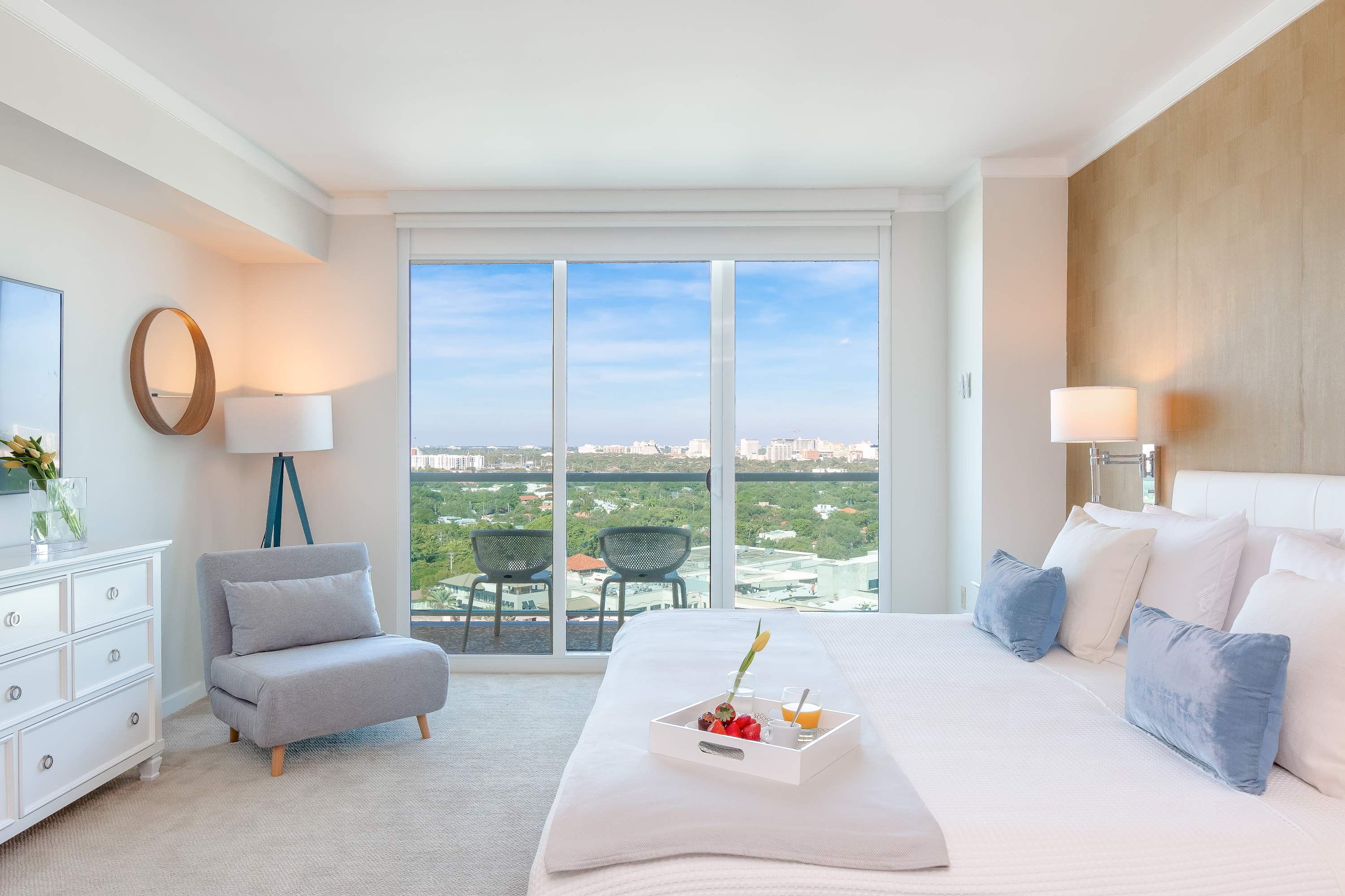 A spacious studio bedroom featuring a king-sized bed with white bedding and blue pillows, a grey accent chair, and a large sliding glass door leading to a balcony with city views. The room has light walls, carpeted flooring, and a textured accent wall.